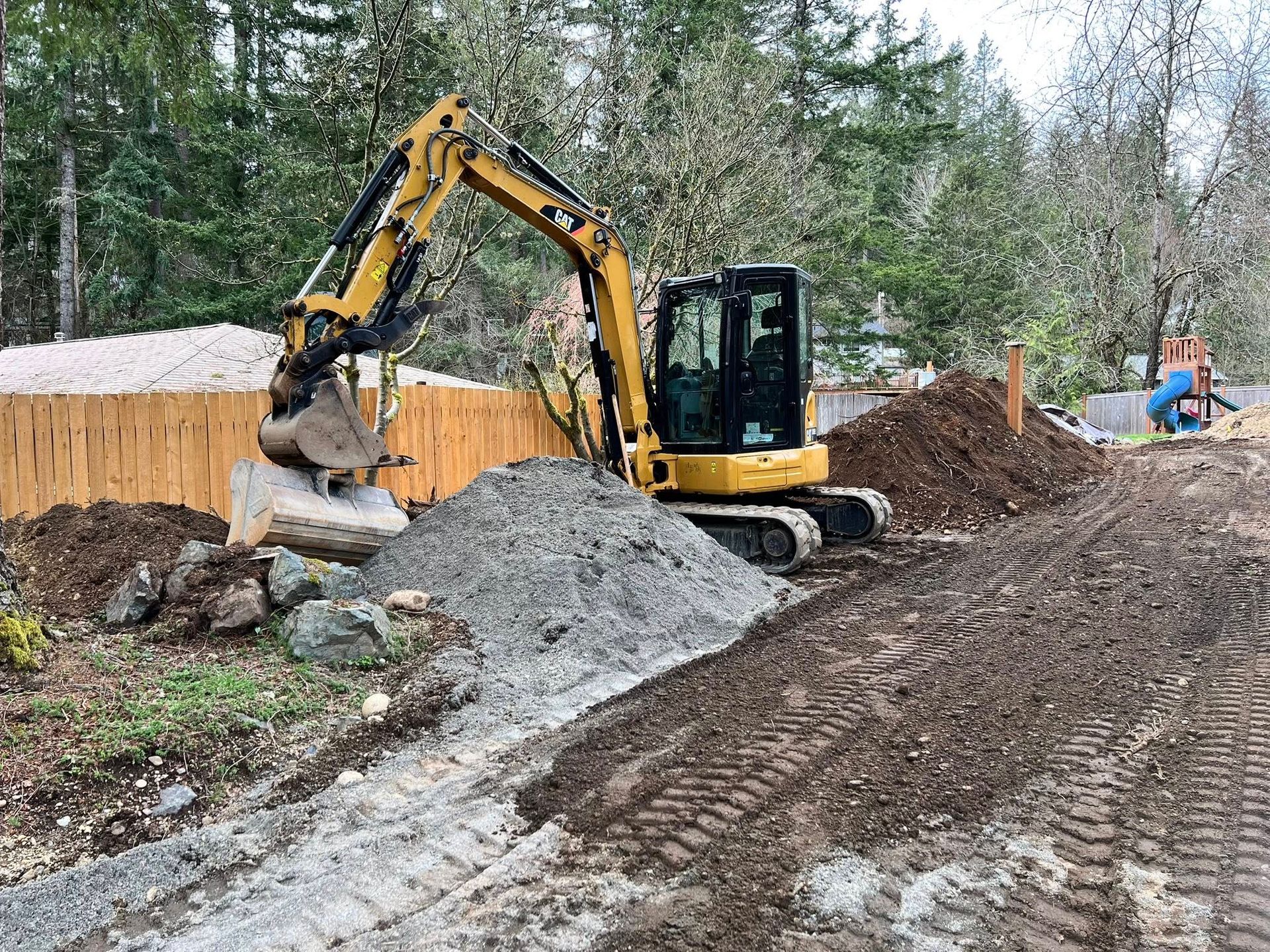 A yellow mini excavator sits on a dirt construction site with a pile of gravel in front of a wooden fence.