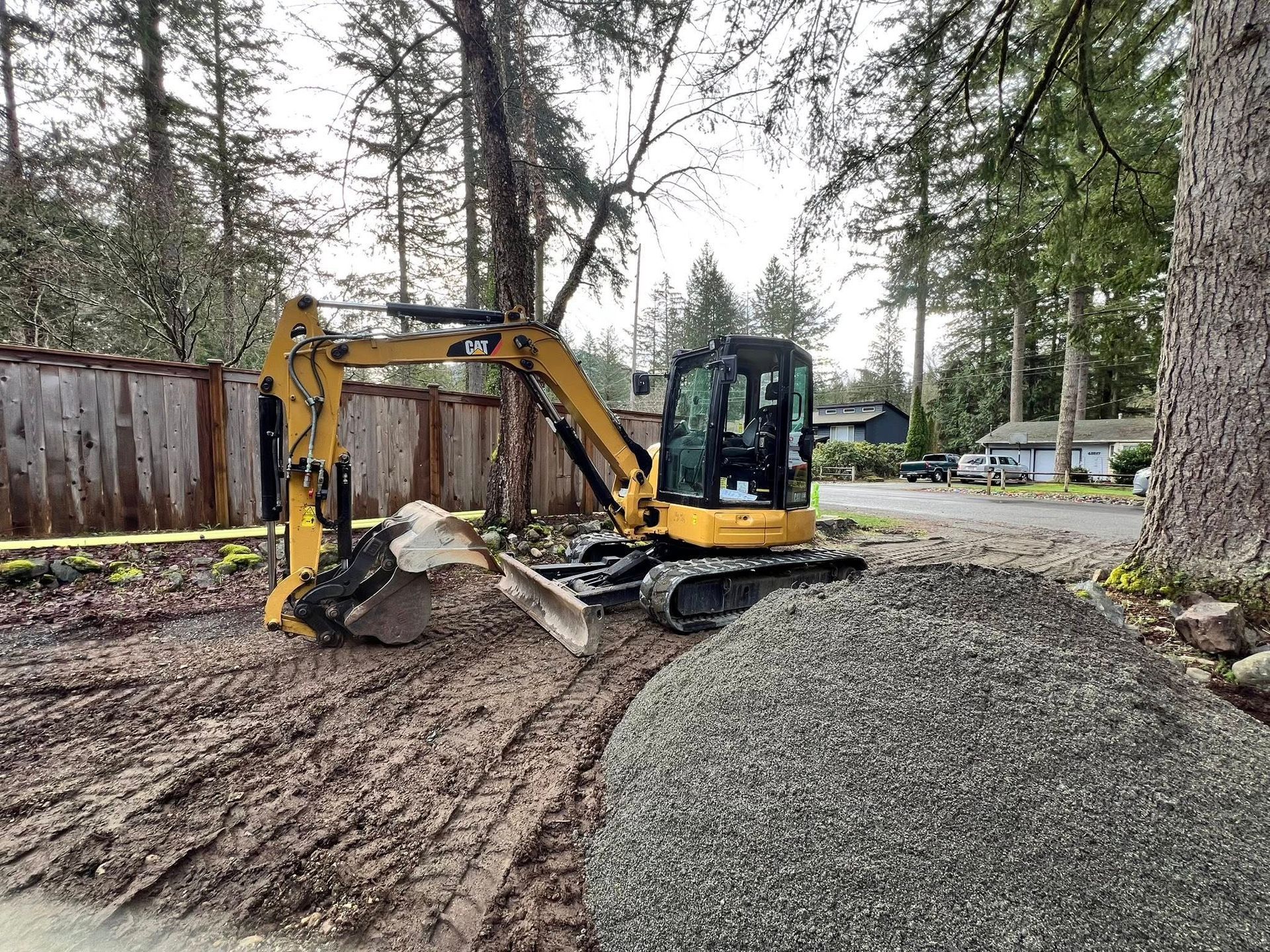 A yellow compact excavator parked on a muddy residential lot next to a large pile of gravel near a wooden fence.