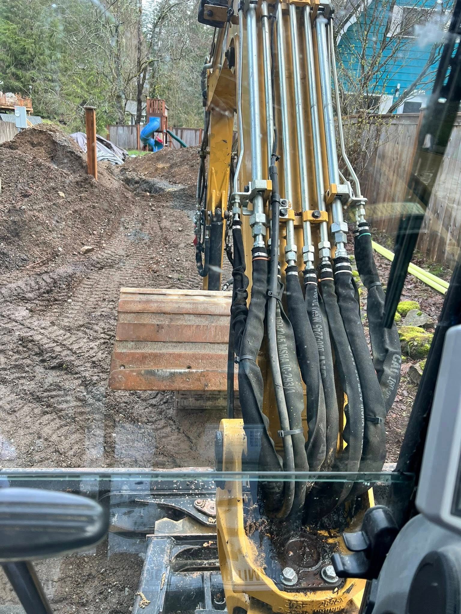 View from an excavator cab showing its yellow mechanical arm, hydraulic hoses, and tracks on a muddy construction site.