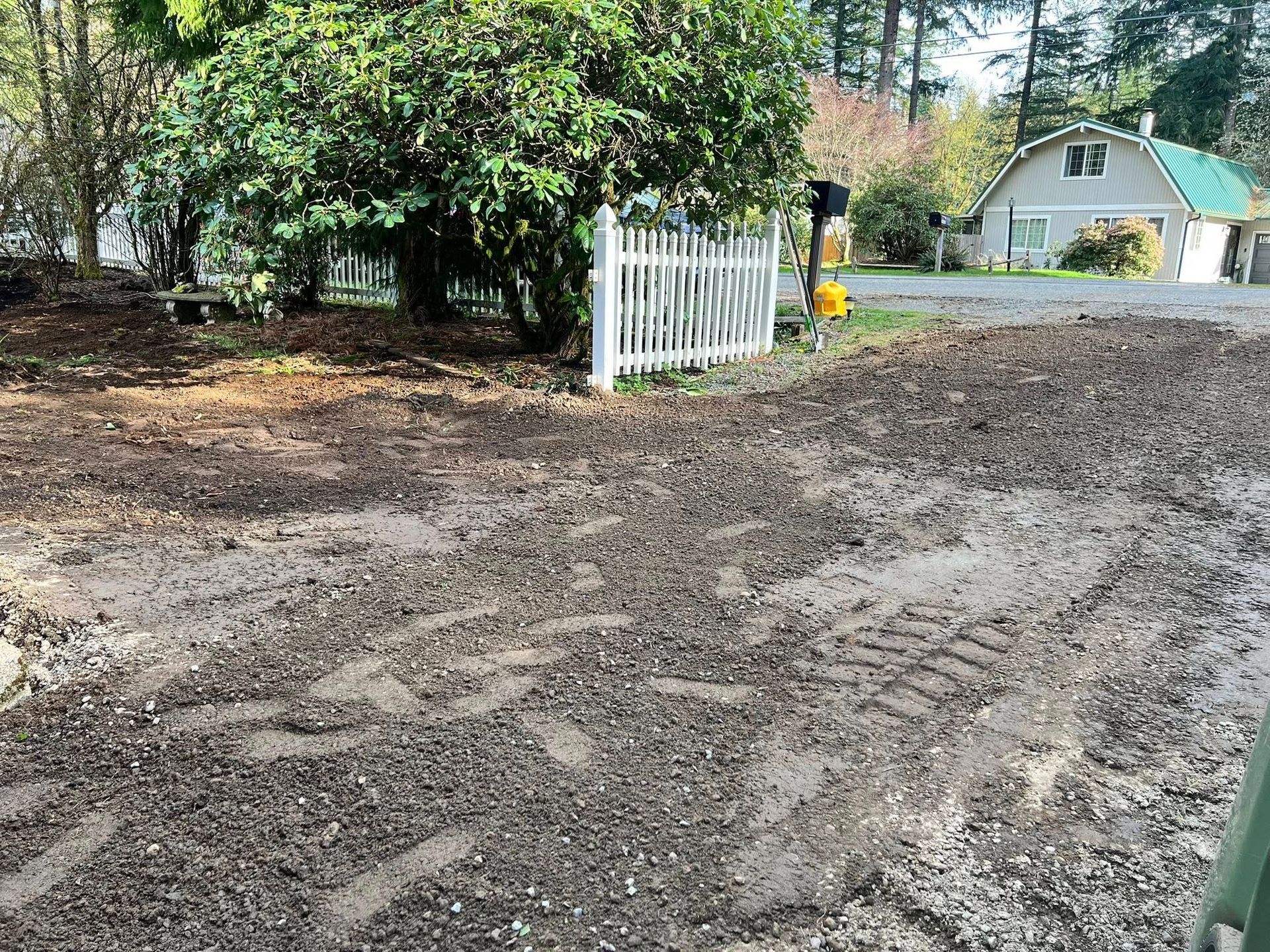 A dirt and gravel driveway leads to a white picket fence, a large bush, and a house in the background.
