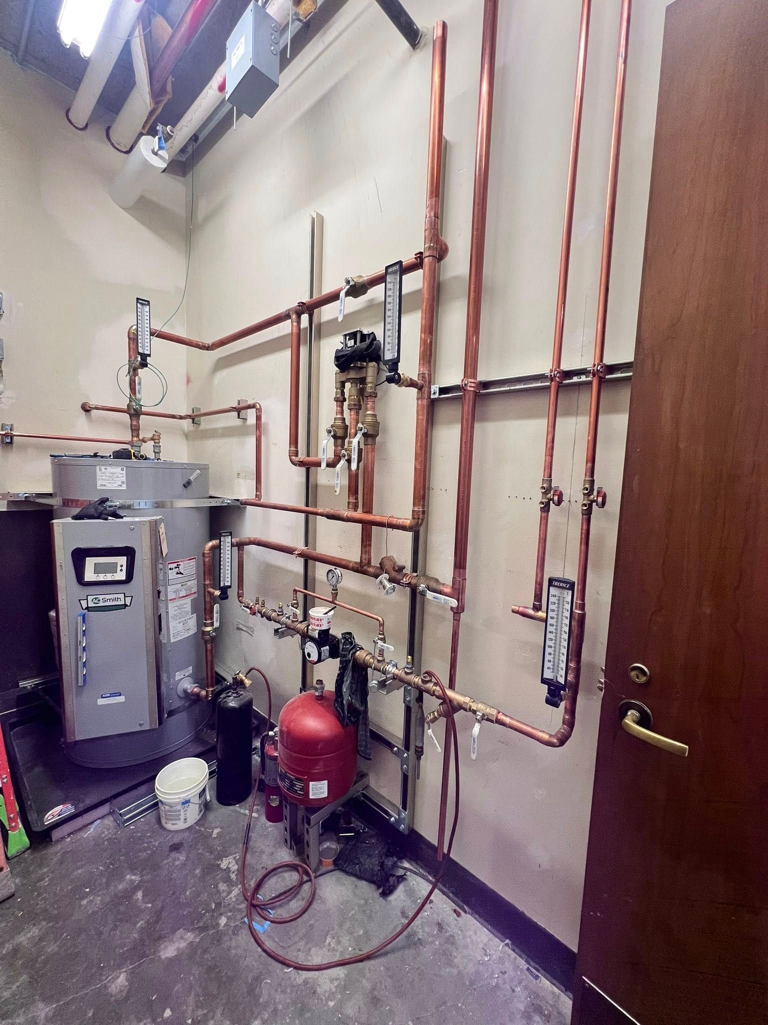 Utility room with a commercial water heater, copper plumbing pipes, valves, and a red expansion tank against a wall.