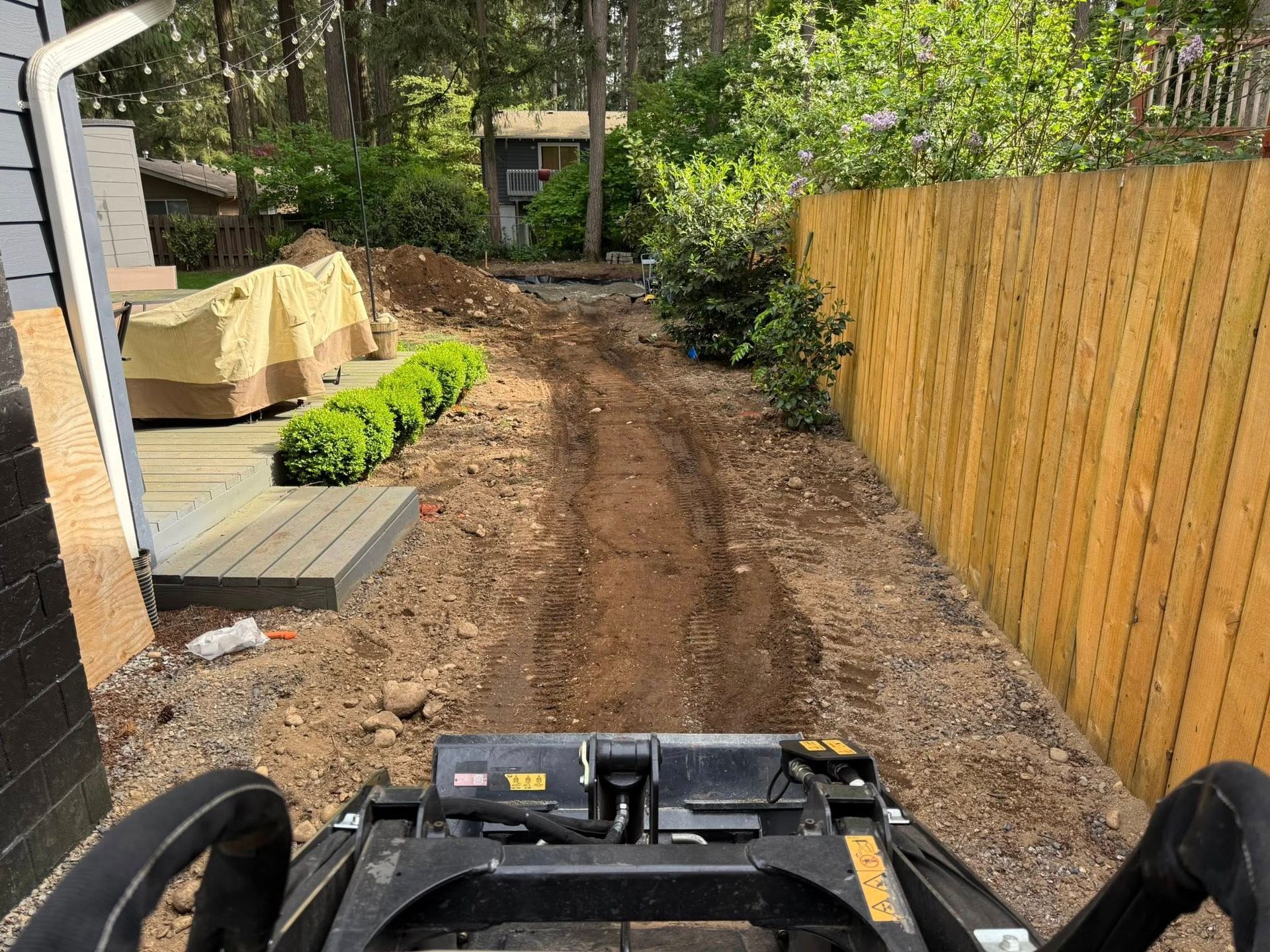 First-person view from a skid steer looking down a dirt trench in a residential yard beside a wooden fence.
