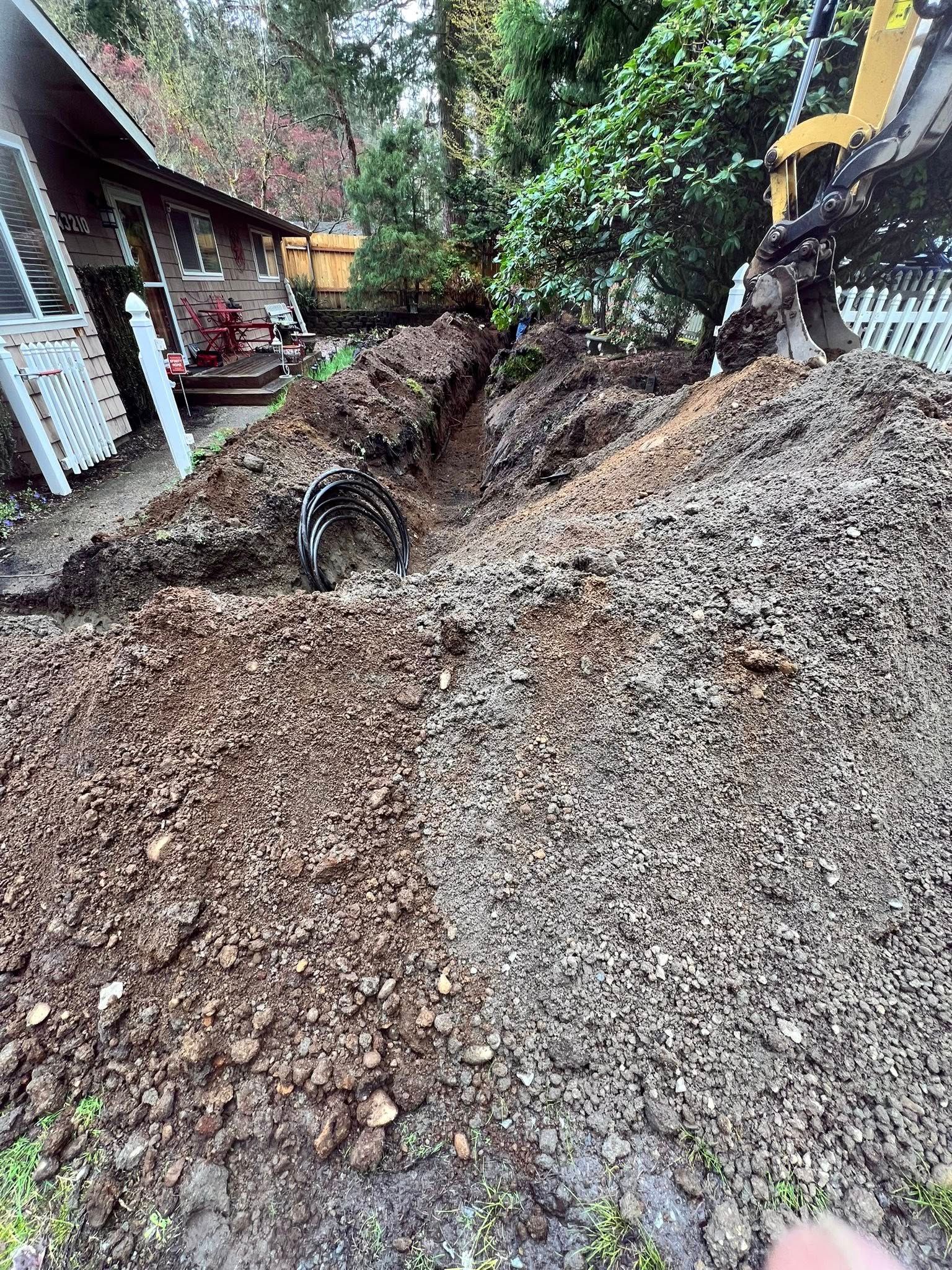 An excavator digs a trench along the side of a house, leaving a large pile of dirt and gravel in the foreground.
