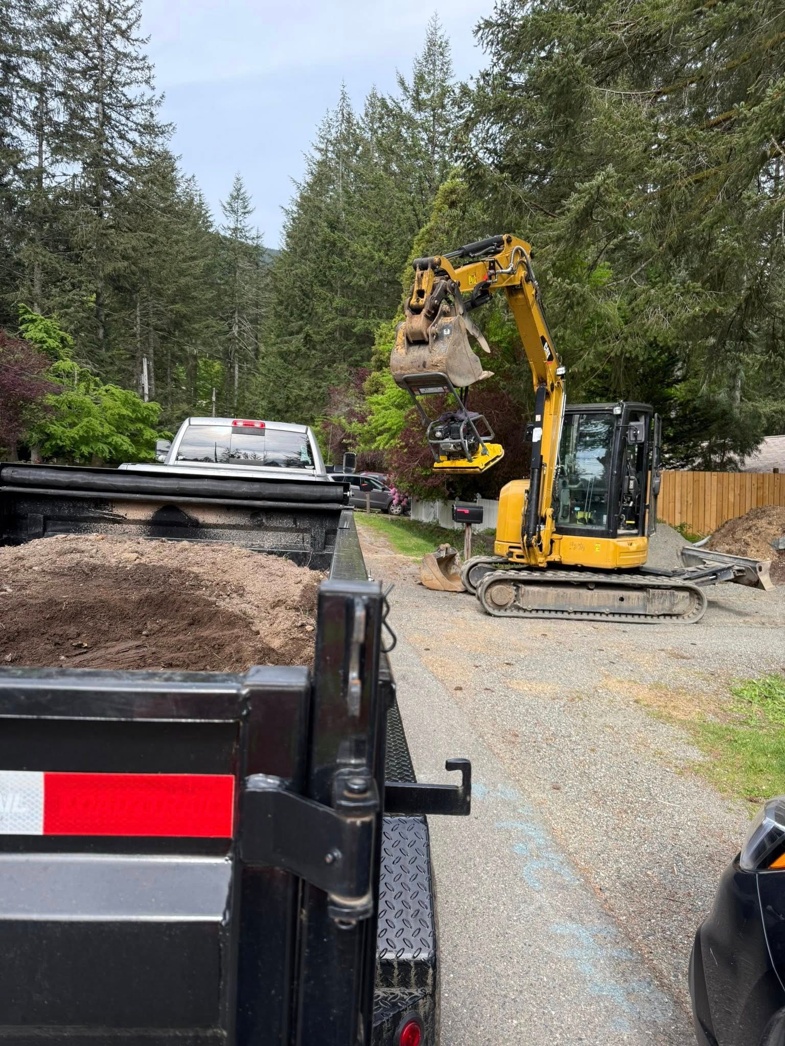 A yellow excavator with an open bucket stands on a gravel driveway, parked next to a truck filled with soil.