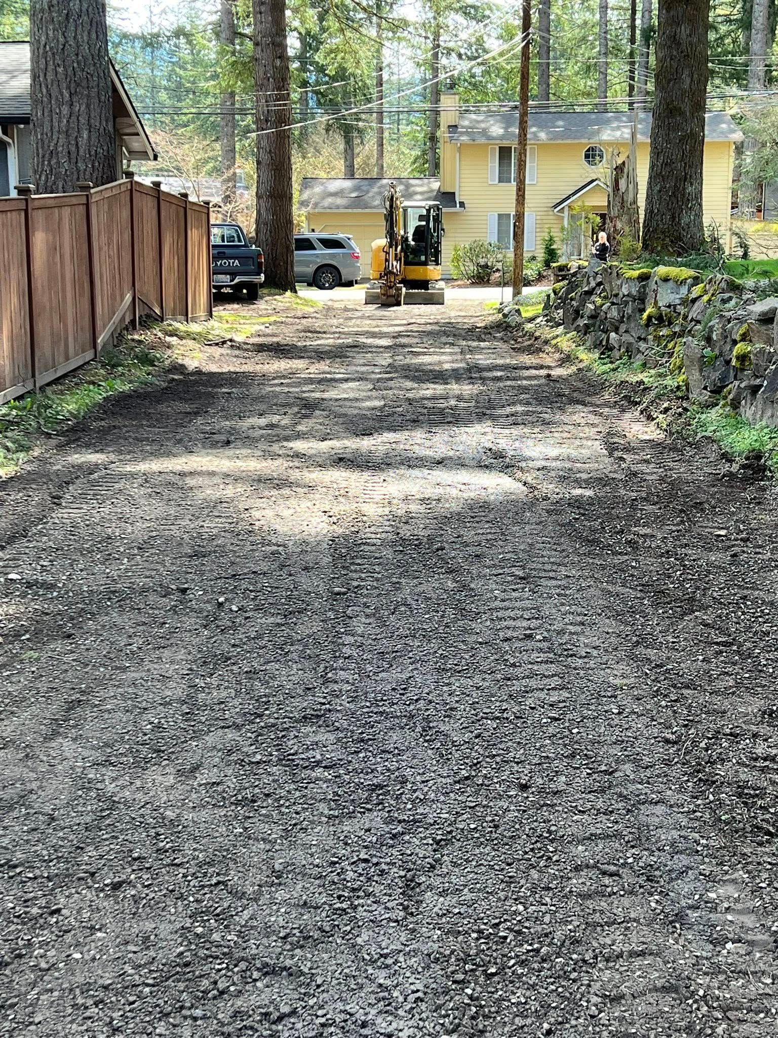 A gravel driveway leading toward a yellow house with a small yellow excavator parked near the entrance.