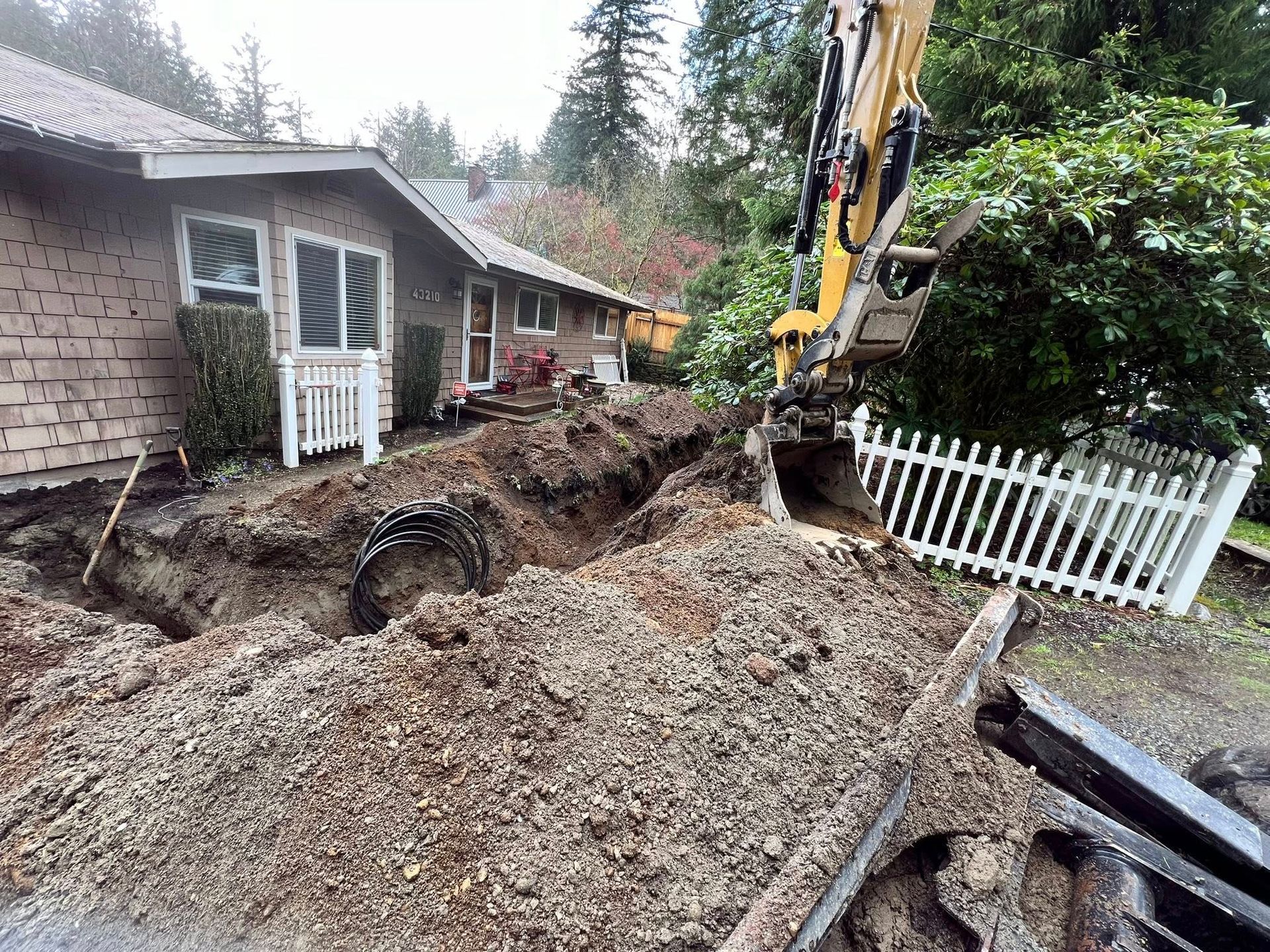 An excavator digs a long trench in a residential yard alongside a house with a white picket fence.