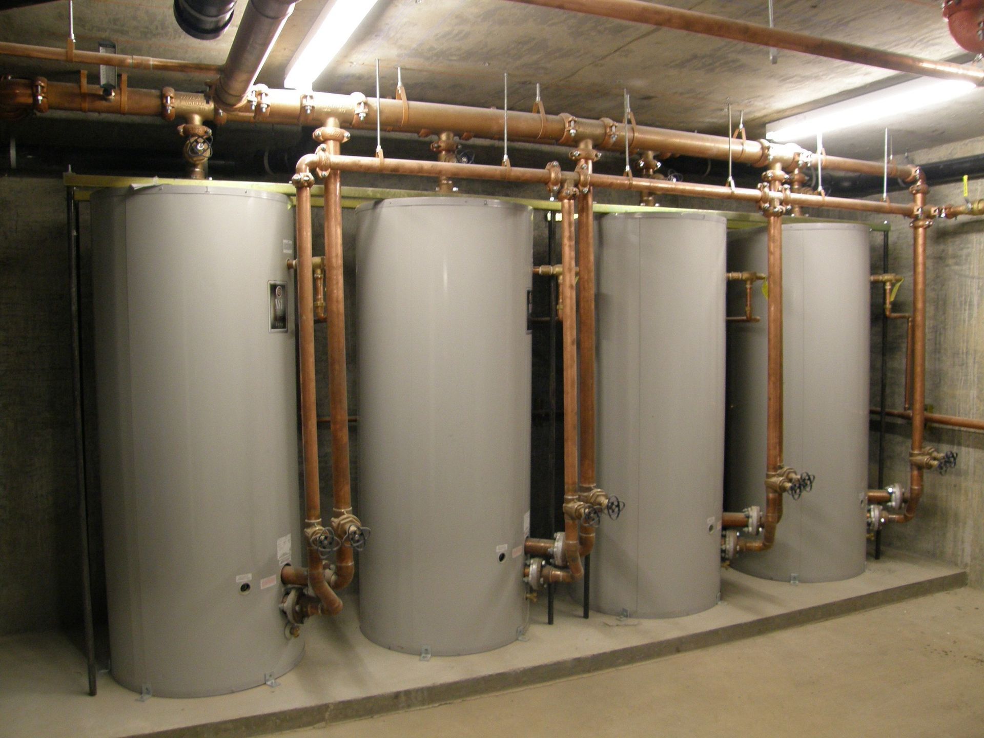 Four grey industrial water heater tanks connected by copper piping in a basement utility room.