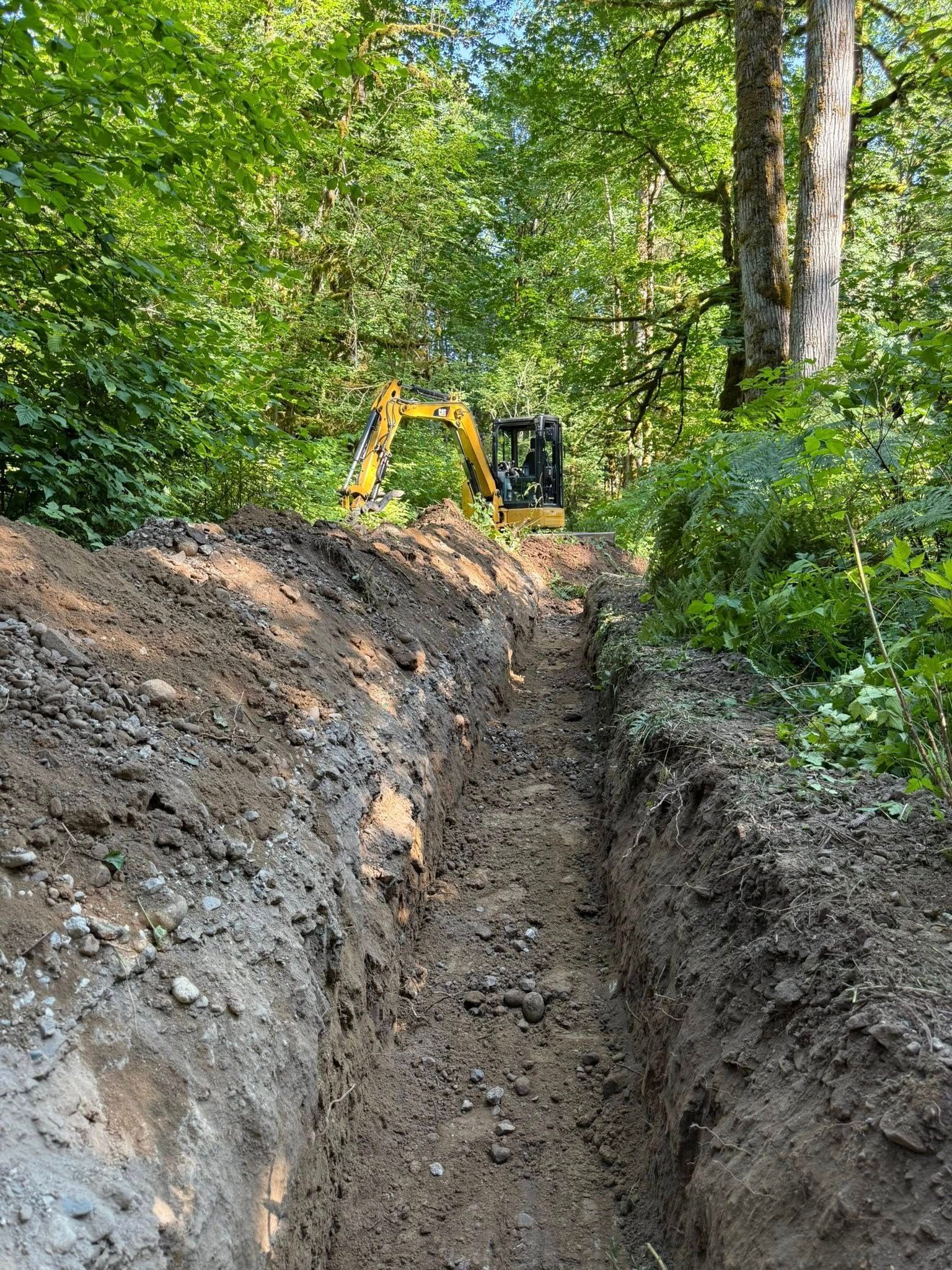 A yellow excavator sits in a wooded area at the end of a long, freshly dug dirt trench.