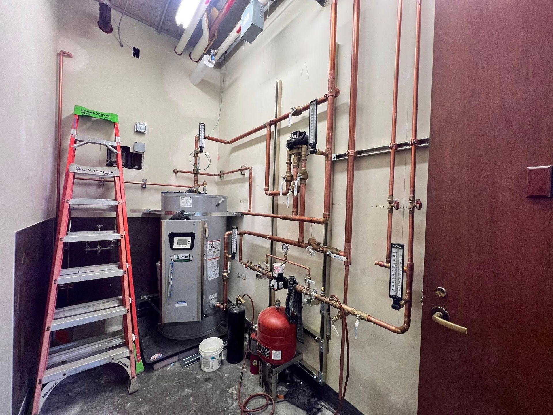 Mechanical room with a water heater, red expansion tank, and copper plumbing pipes on the wall next to a red ladder.