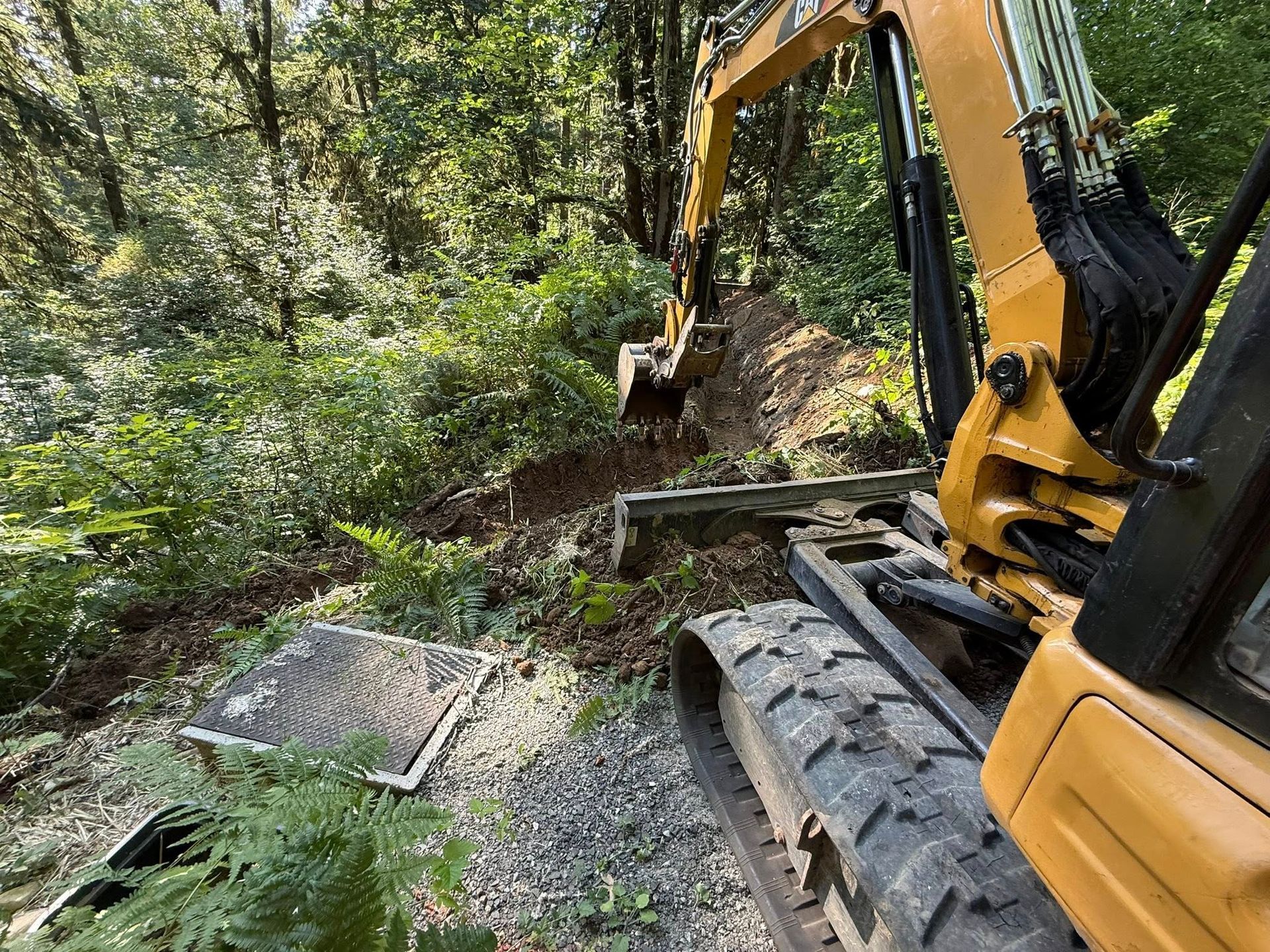 A yellow excavator sits in a wooded area, digging a trench next to a square metal utility cover.