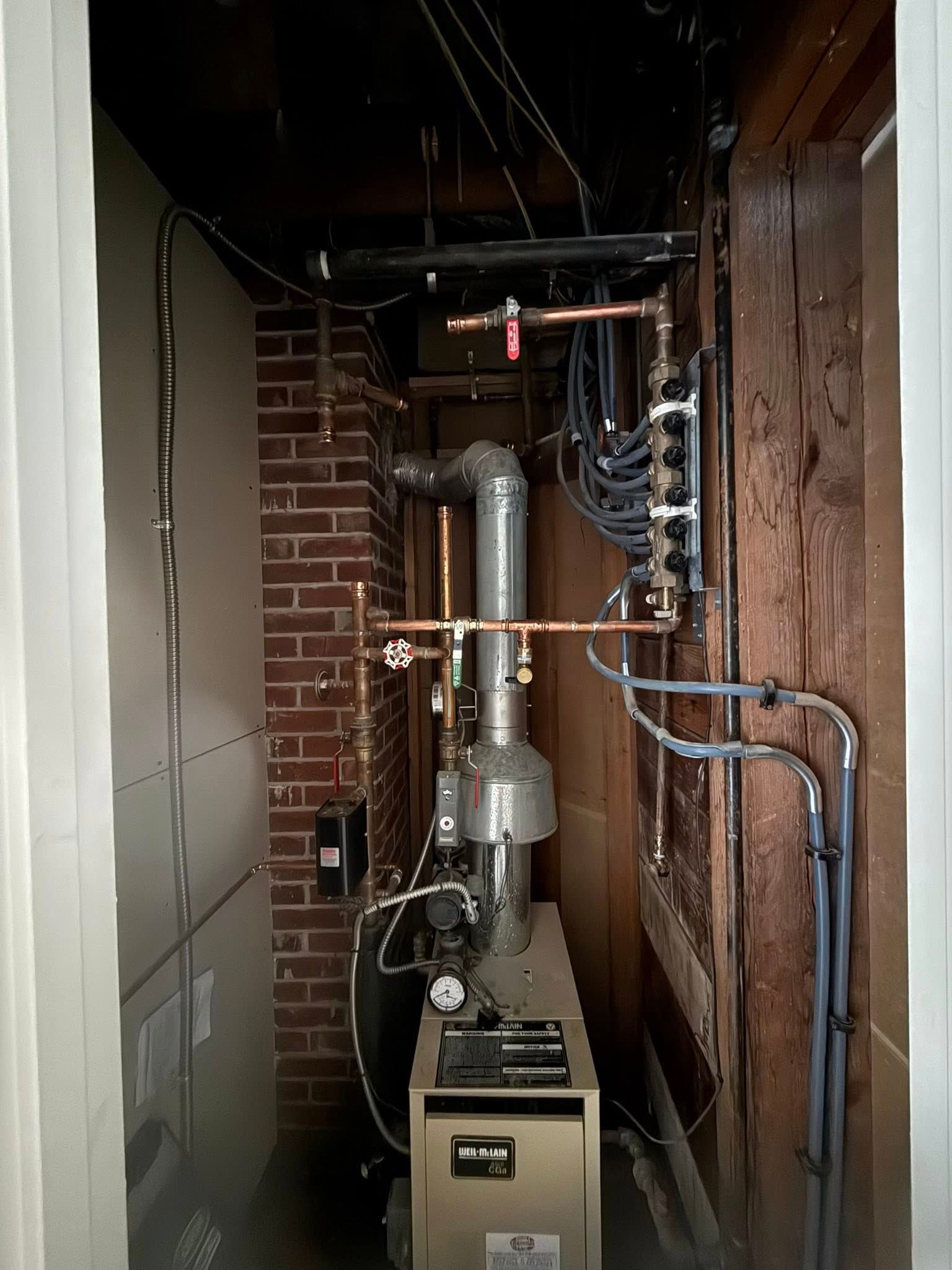 A furnace in a narrow utility space with visible brick, copper piping, and a metal exhaust vent.