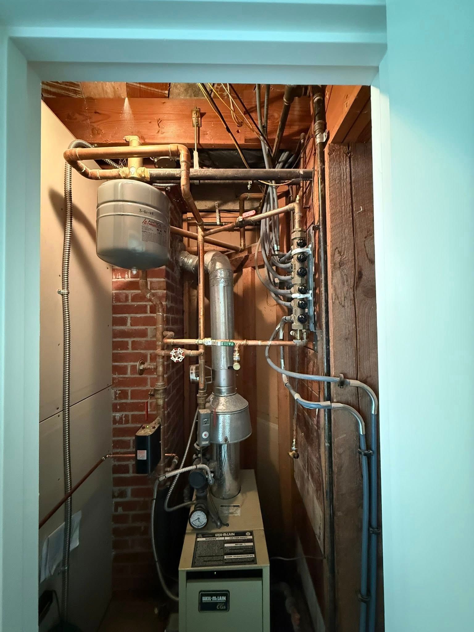 A mechanical closet featuring a boiler, copper pipes, a gray expansion tank, and a metal exhaust vent against brick walls.