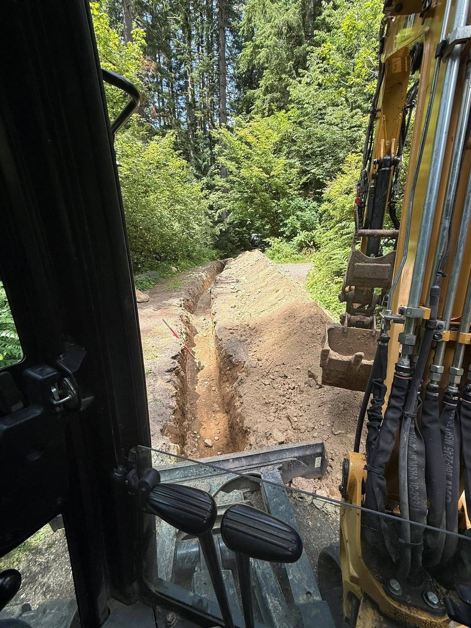 View from inside an excavator cab looking out at a trench dug into dirt with lush green trees in the background.