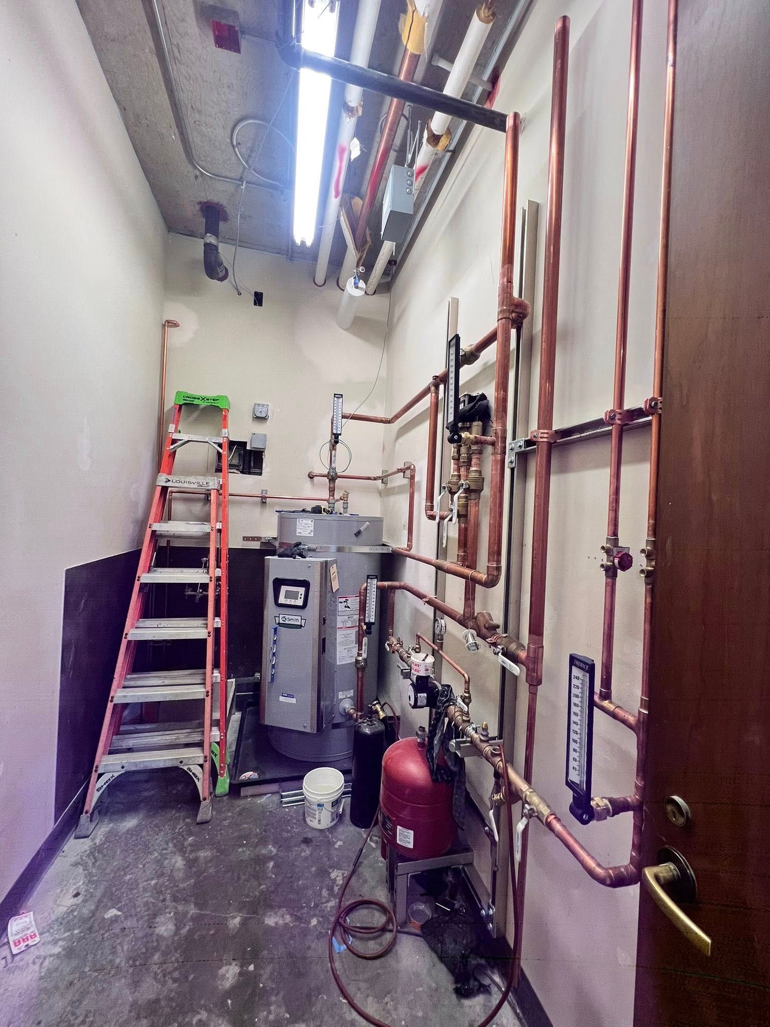 A utility closet showing a water heater, a red expansion tank, and a complex network of copper plumbing pipes.