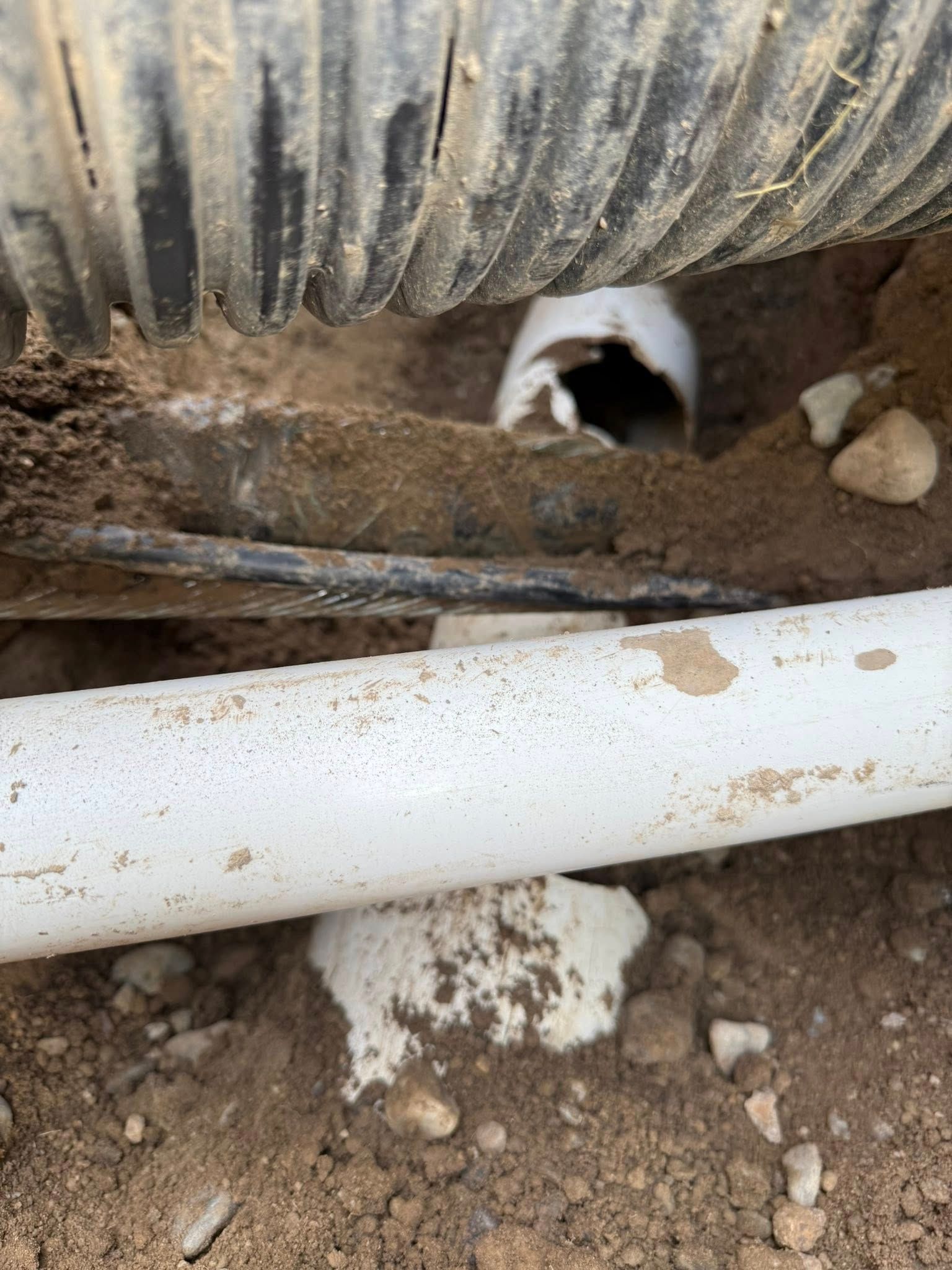 A black corrugated pipe and a white PVC pipe partially exposed in a dirt trench, showing a section of broken pipe.