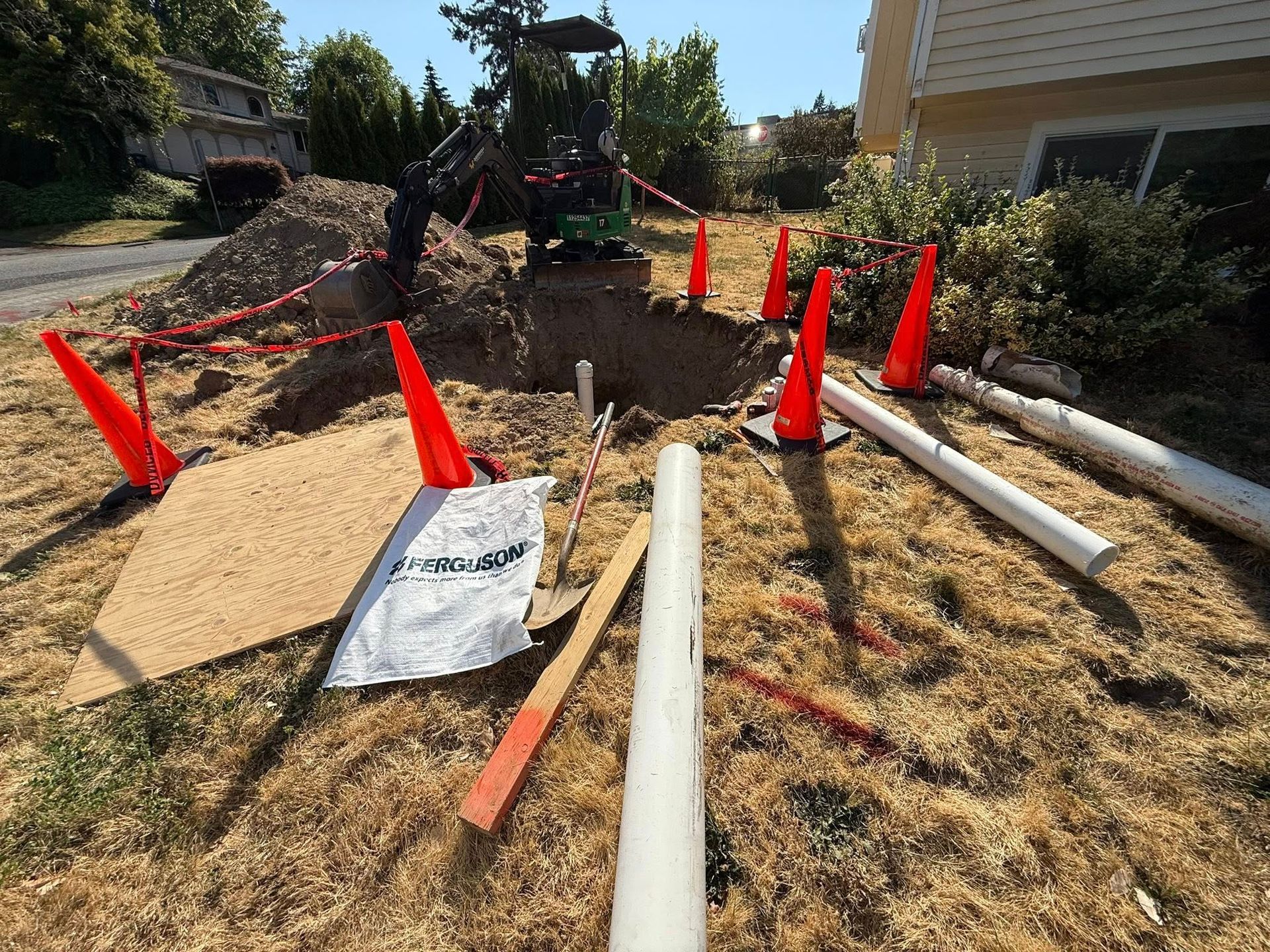A small excavator digs a hole in a residential yard near a house, marked by safety cones and white PVC piping.