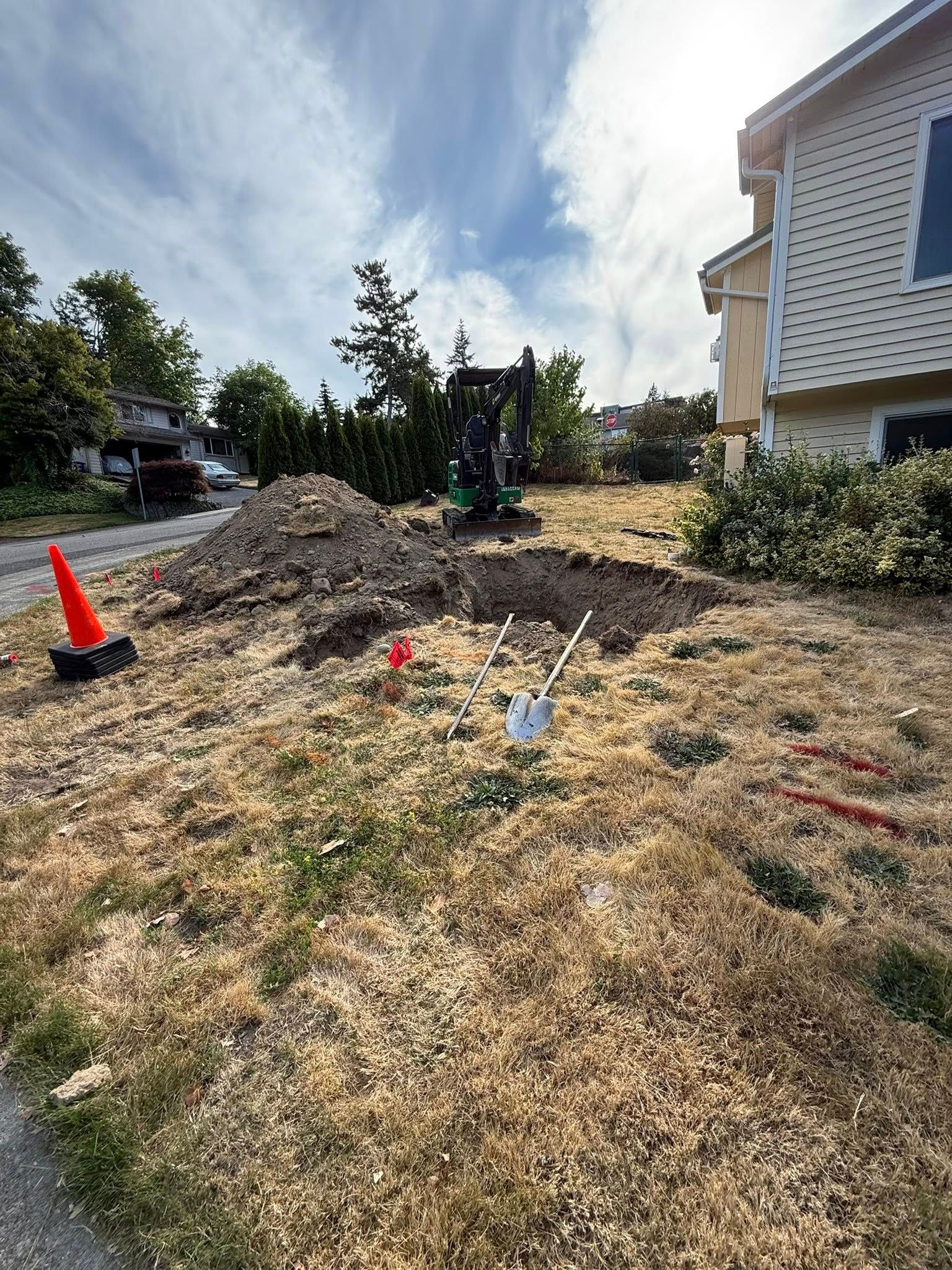 An excavator digs a trench in a front yard next to a house, with a pile of dirt, a safety cone, and a shovel nearby.