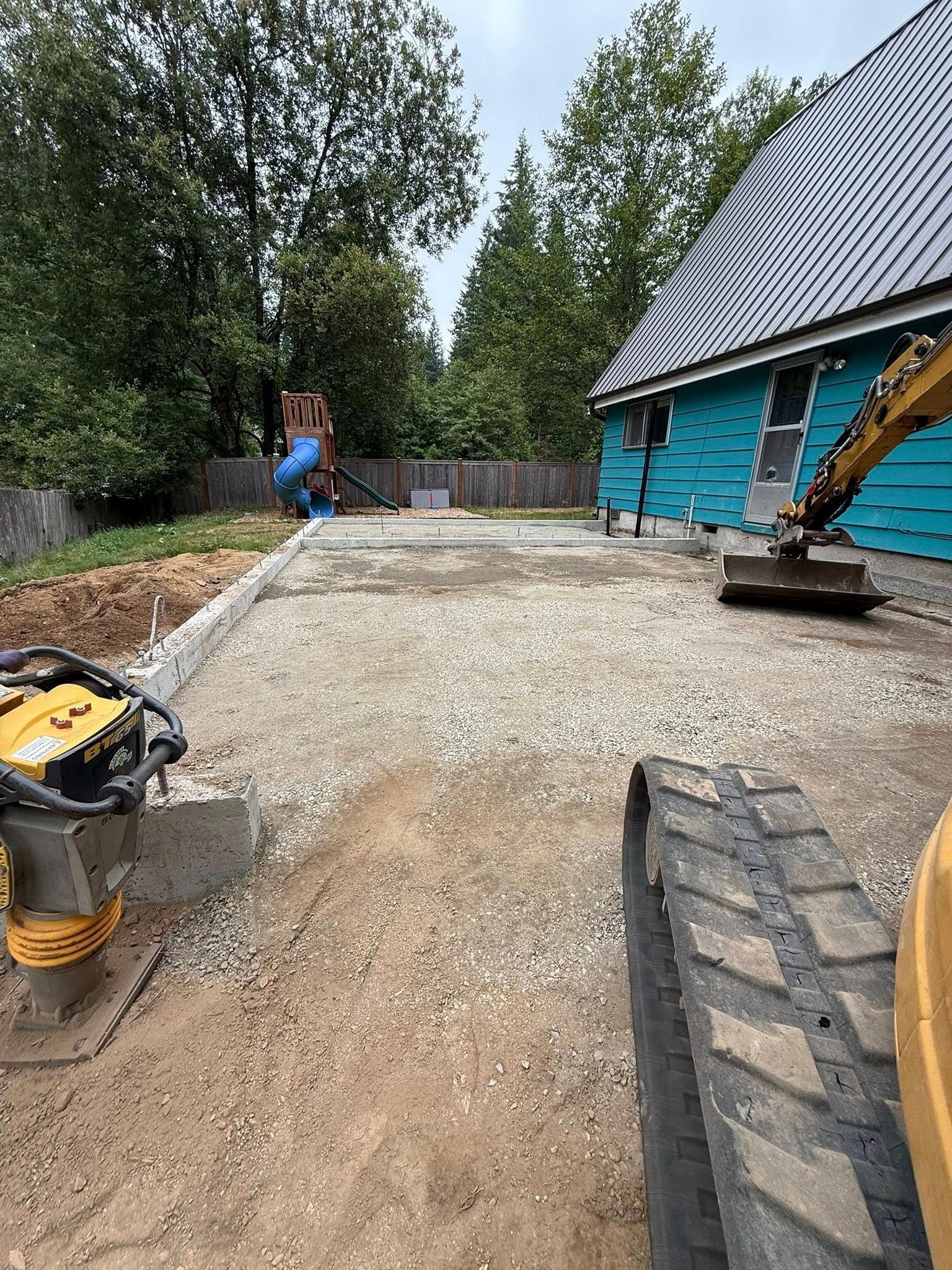 Construction site with gravel ground and a compactor in front of a blue house with construction equipment.