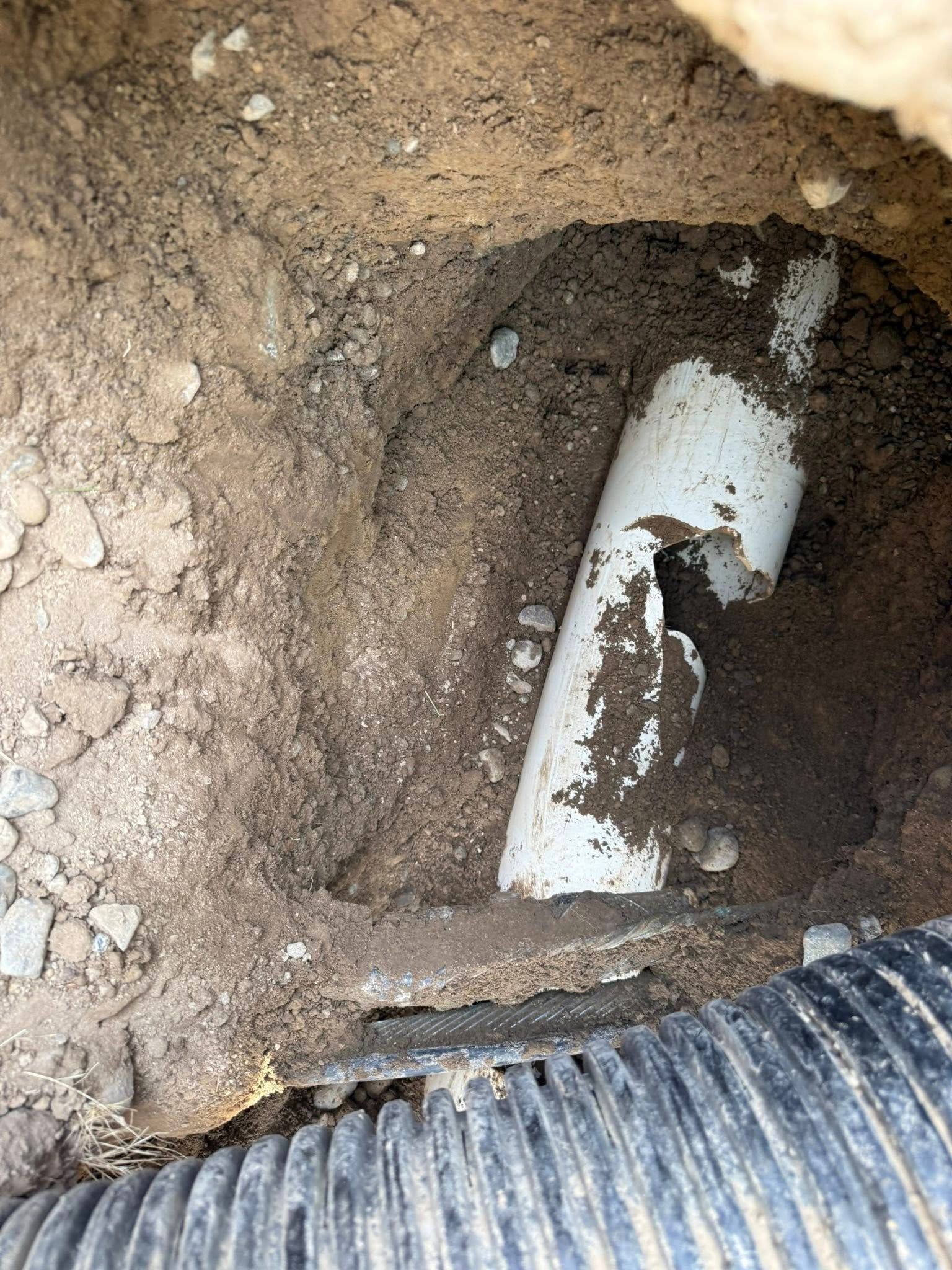 A broken white PVC pipe partially exposed in a dirt trench next to a section of black corrugated drainage pipe.