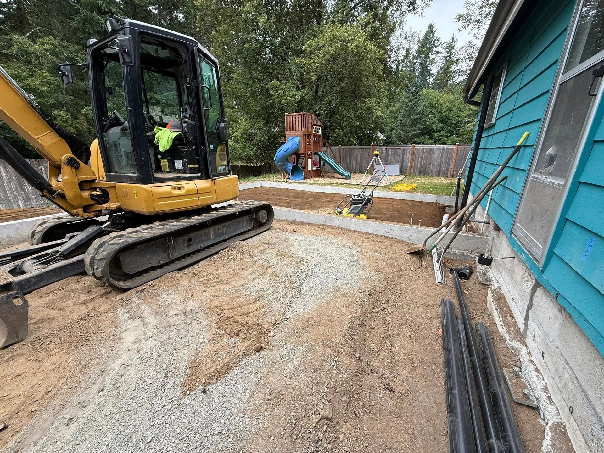 A yellow excavator parked next to a blue house, with a freshly graded gravel area in a backyard near a play set.
