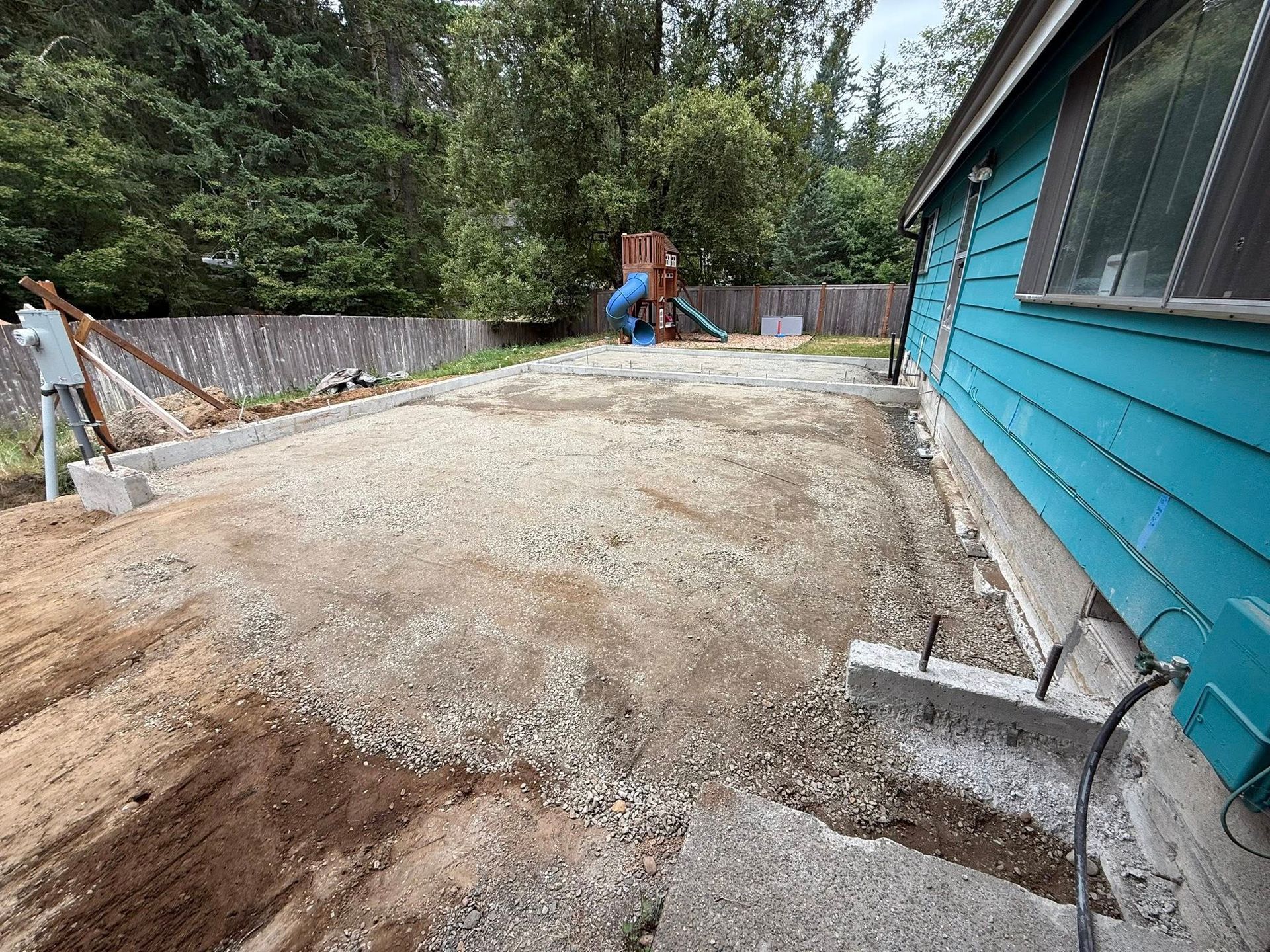 A gravel-covered construction site next to a blue house with concrete footings set for a new foundation.