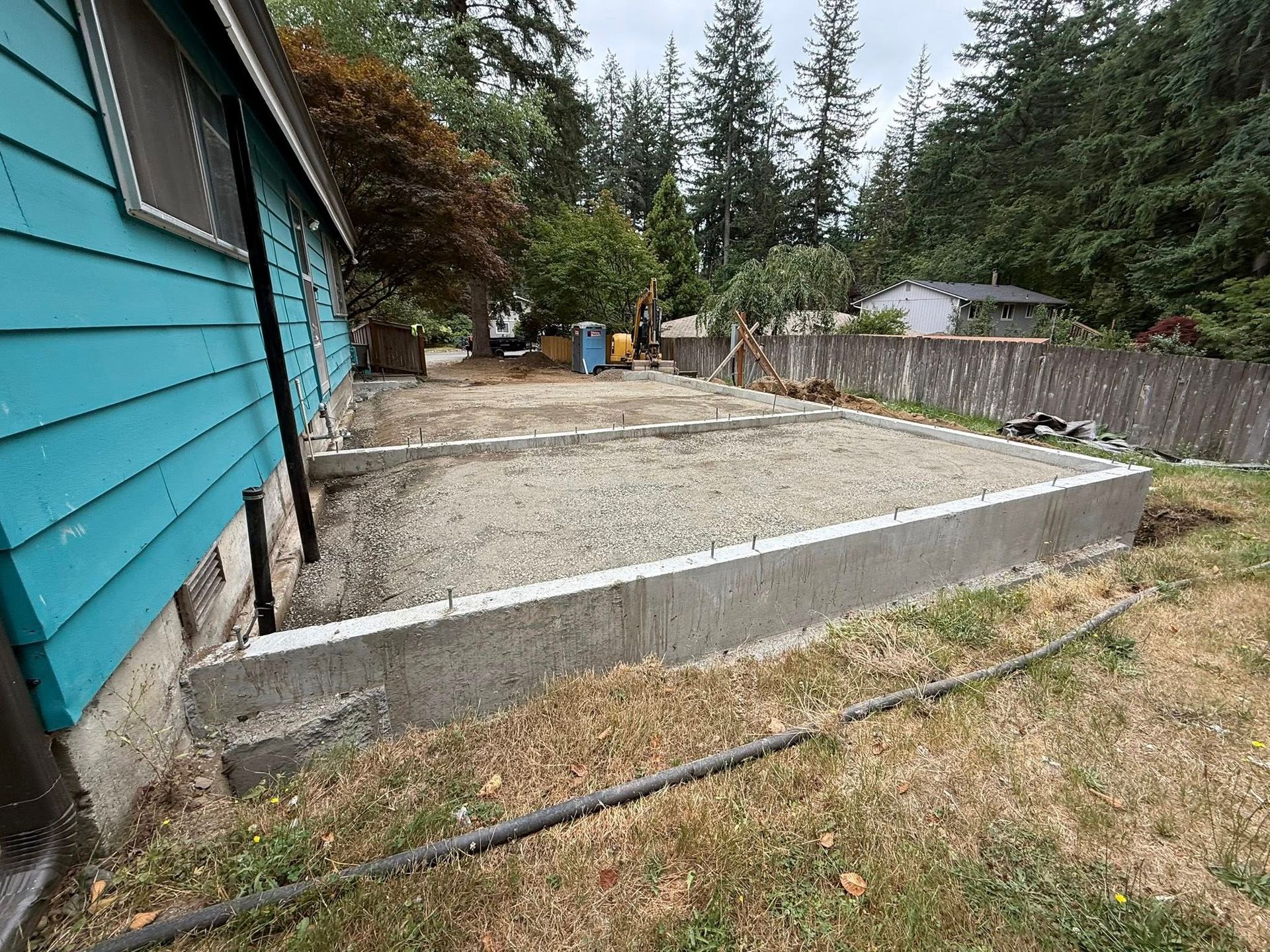 A concrete foundation with a gravel fill sits adjacent to the side of a turquoise house, with a wooden fence in the back.