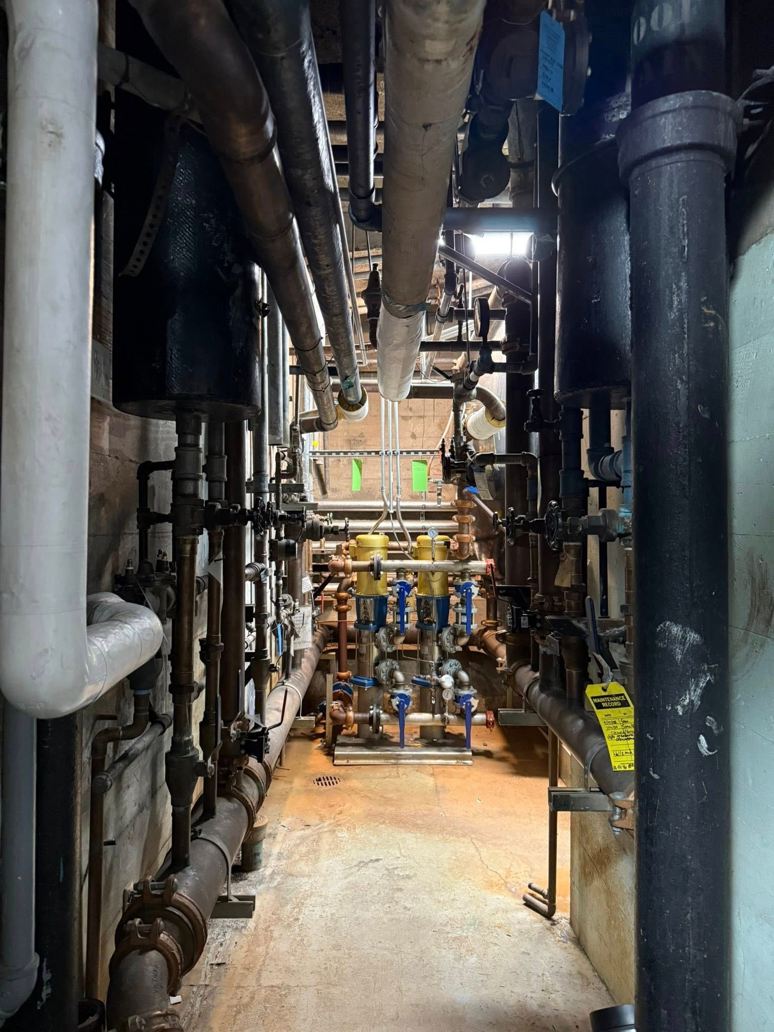 A view into a narrow mechanical utility corridor filled with various pipes, valves, and a light fixture at the end.