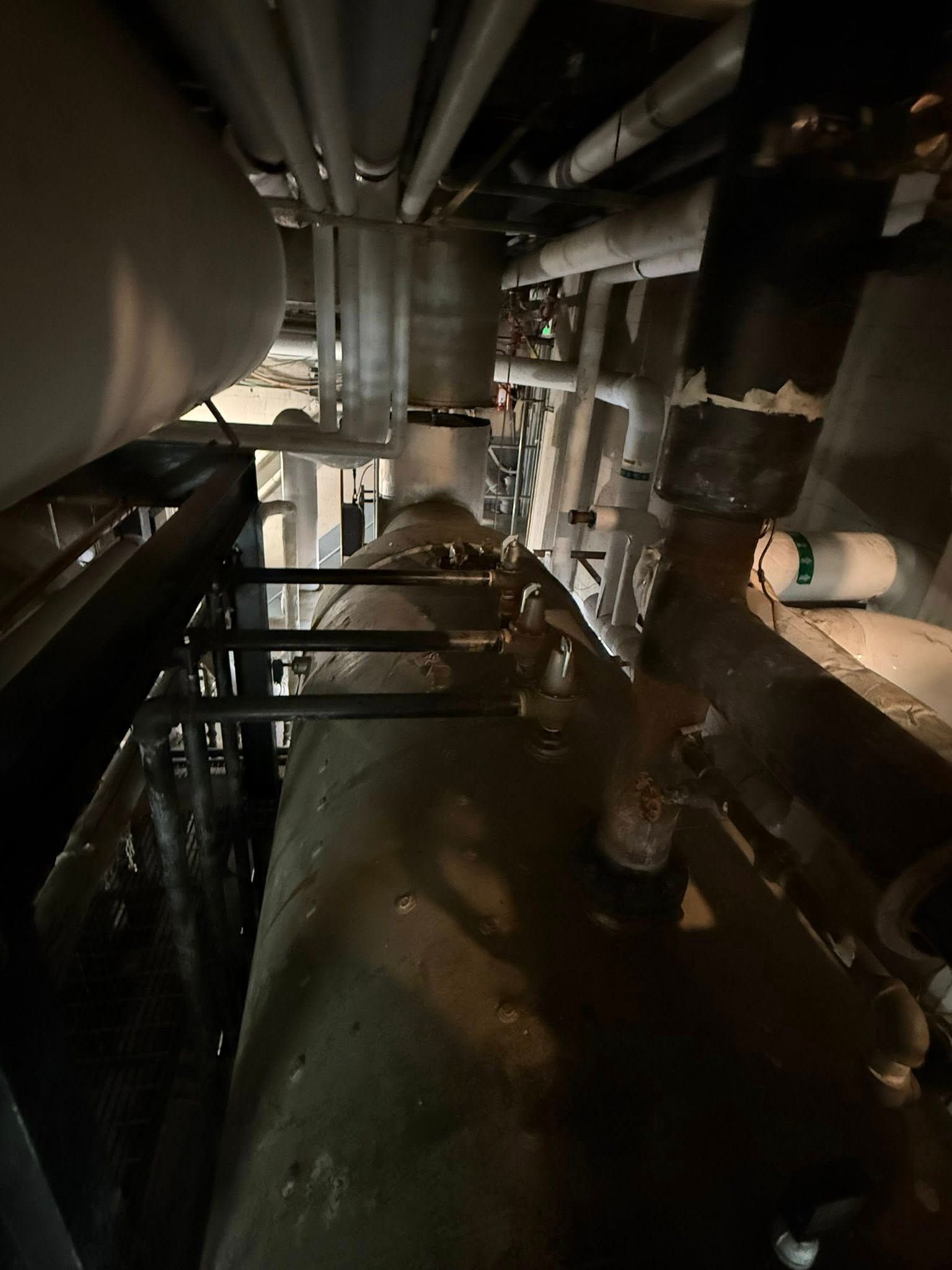 A dark, cluttered view of industrial pipes and mechanical tanks in a dimly lit, narrow maintenance space.