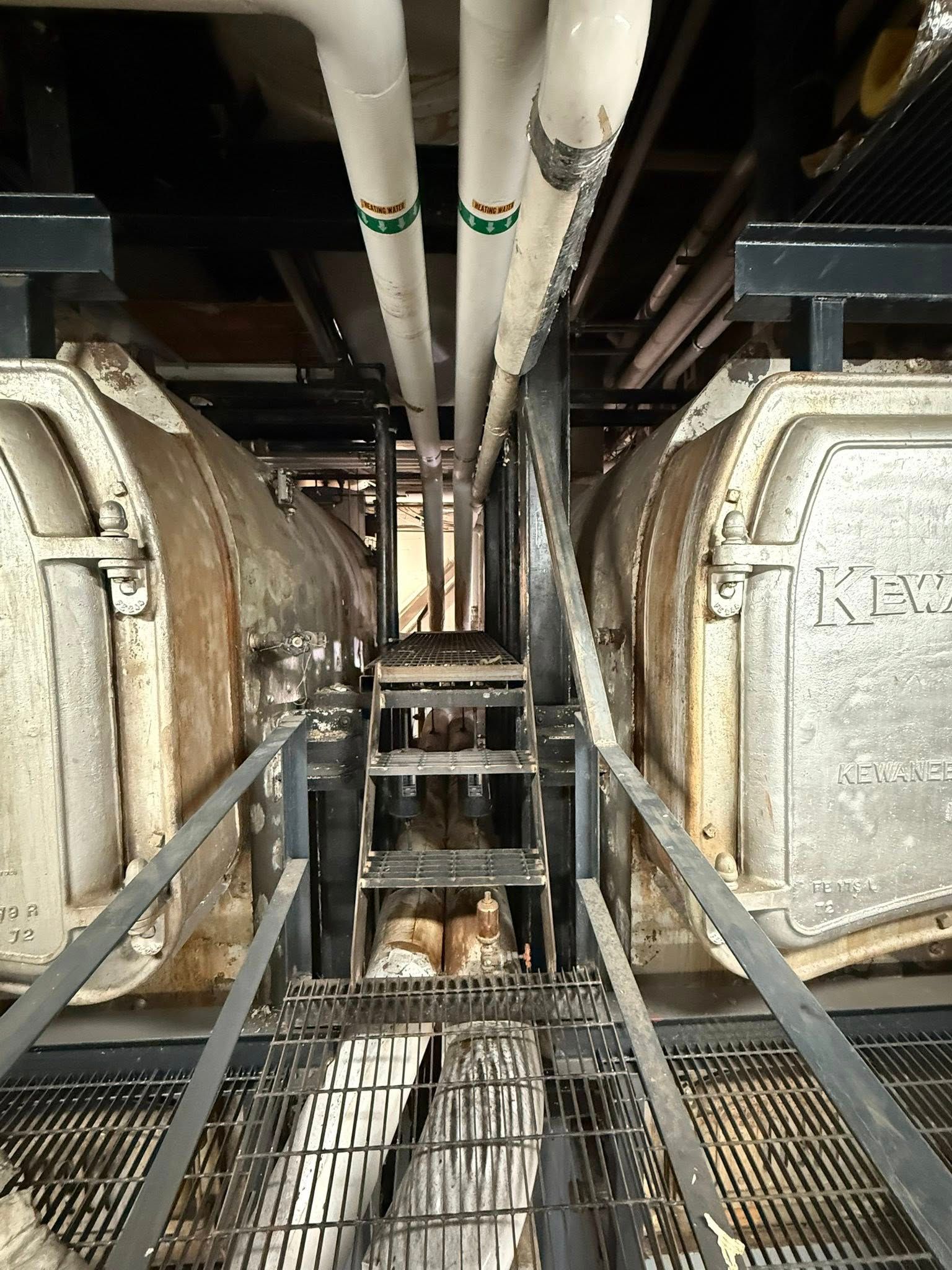 A metal catwalk between two large industrial boilers with white pipes overhead in a dimly lit, industrial mechanical room.