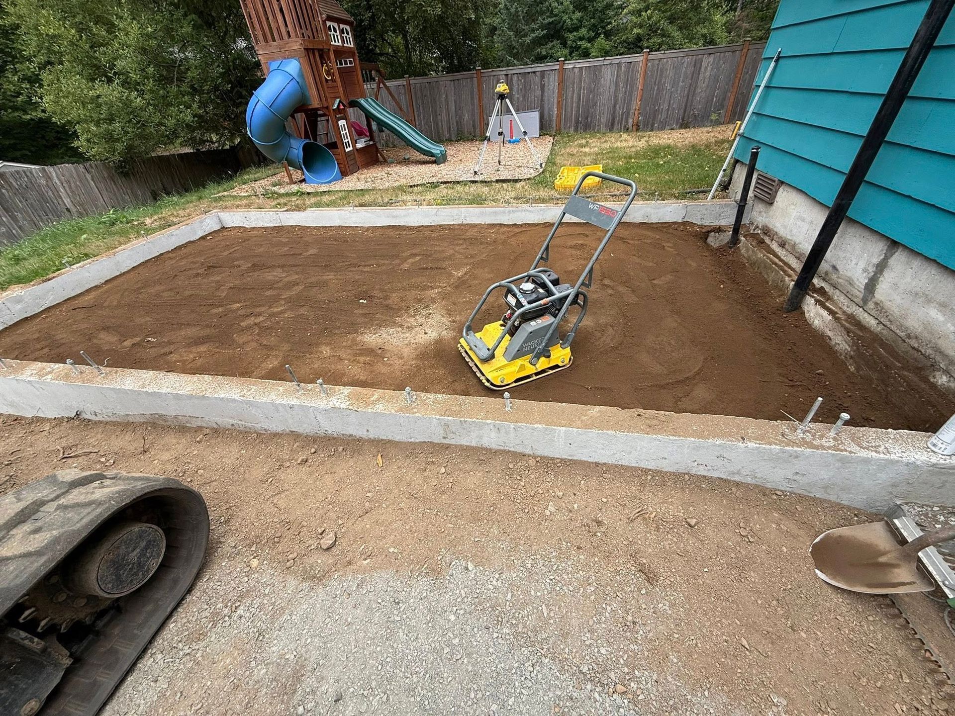 A yellow plate compactor sits in a rectangular dirt area framed by concrete borders, next to a teal house siding.