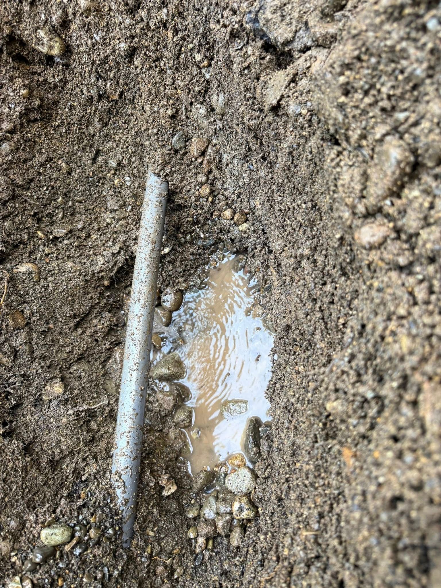 A metal stake or rod stands vertically in a narrow, muddy trench partially filled with water.