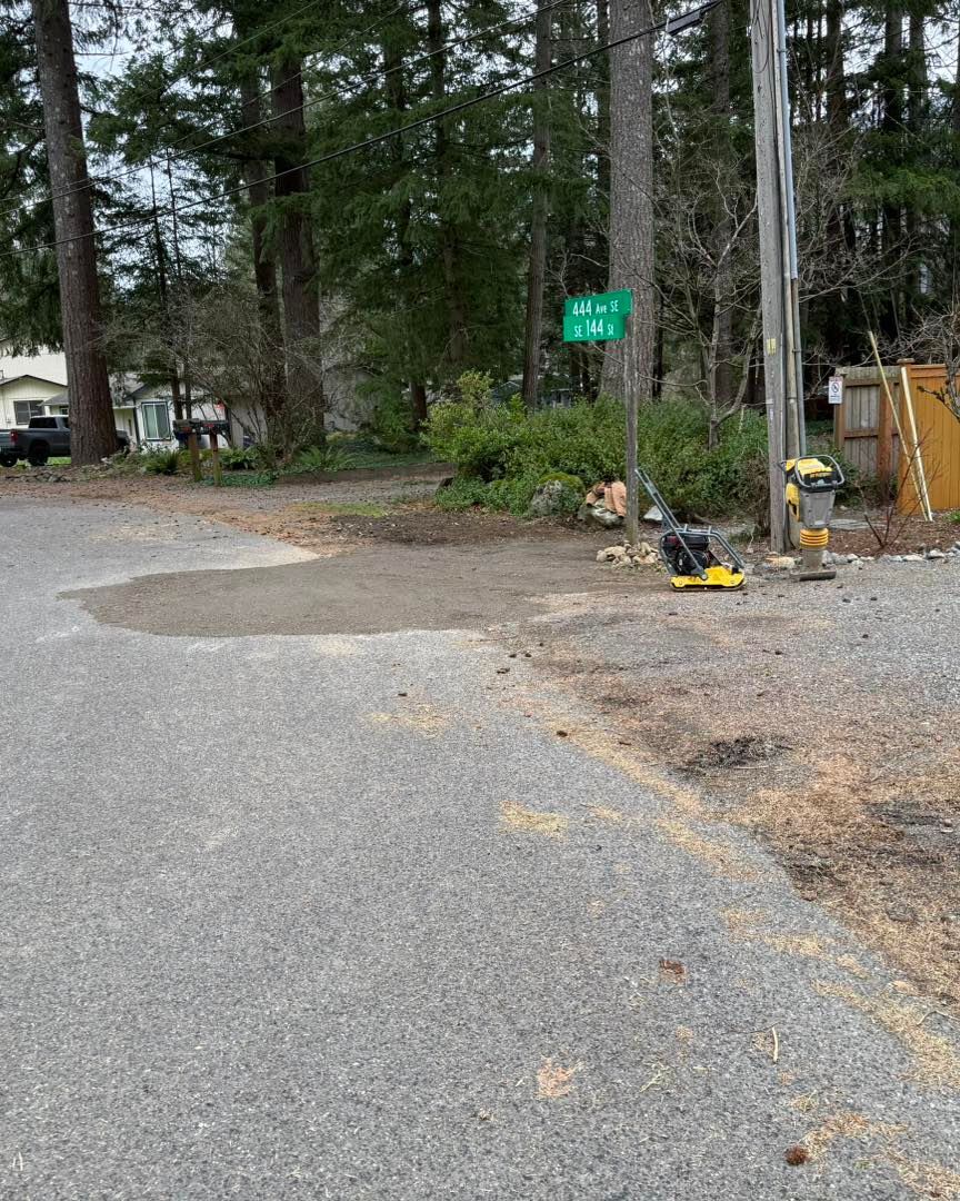 A gravel driveway with a patch of fresh, packed soil and construction equipment near a green street sign in a wooded area.