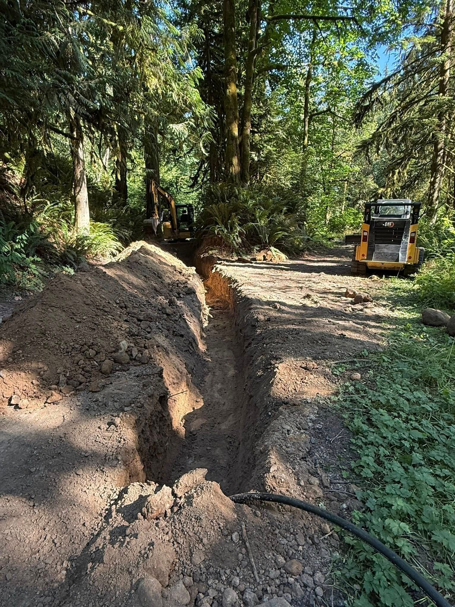 A trench dug in a wooded area with an excavator and a skid steer parked in the background.