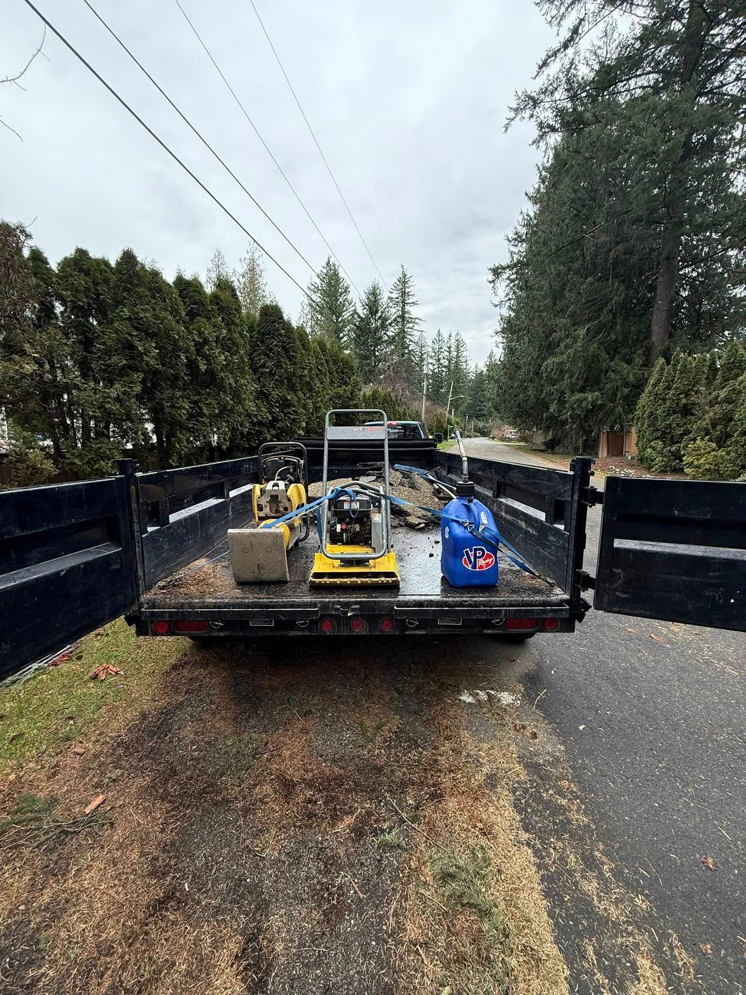 A dump trailer loaded with equipment and a blue fuel jug, parked on a road with trees in the background.