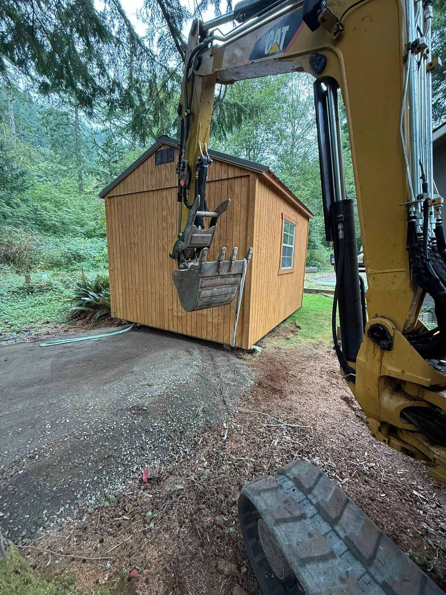 An excavator bucket hangs suspended in front of a wooden shed in a wooded area.