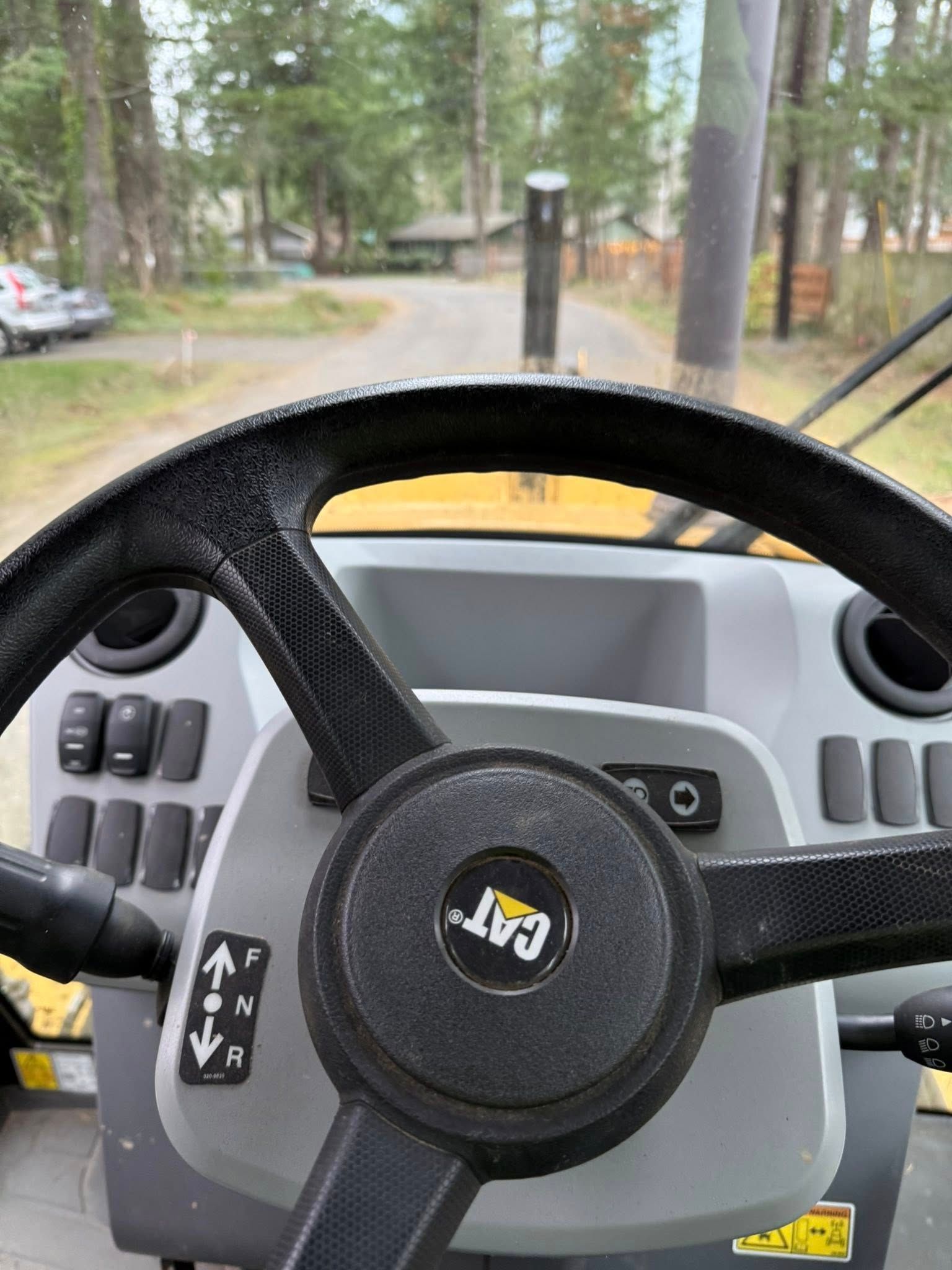 First-person view from the steering wheel of a yellow Caterpillar construction vehicle on a road near a wooded area.