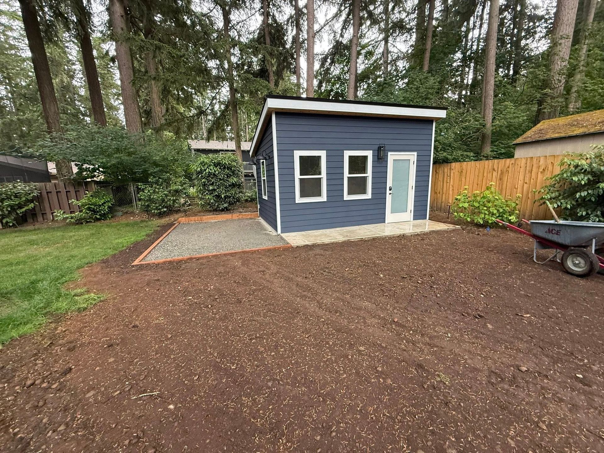 A blue shed sits on a gravel base in a backyard next to a brown wooden fence, with surrounding trees and bare soil.