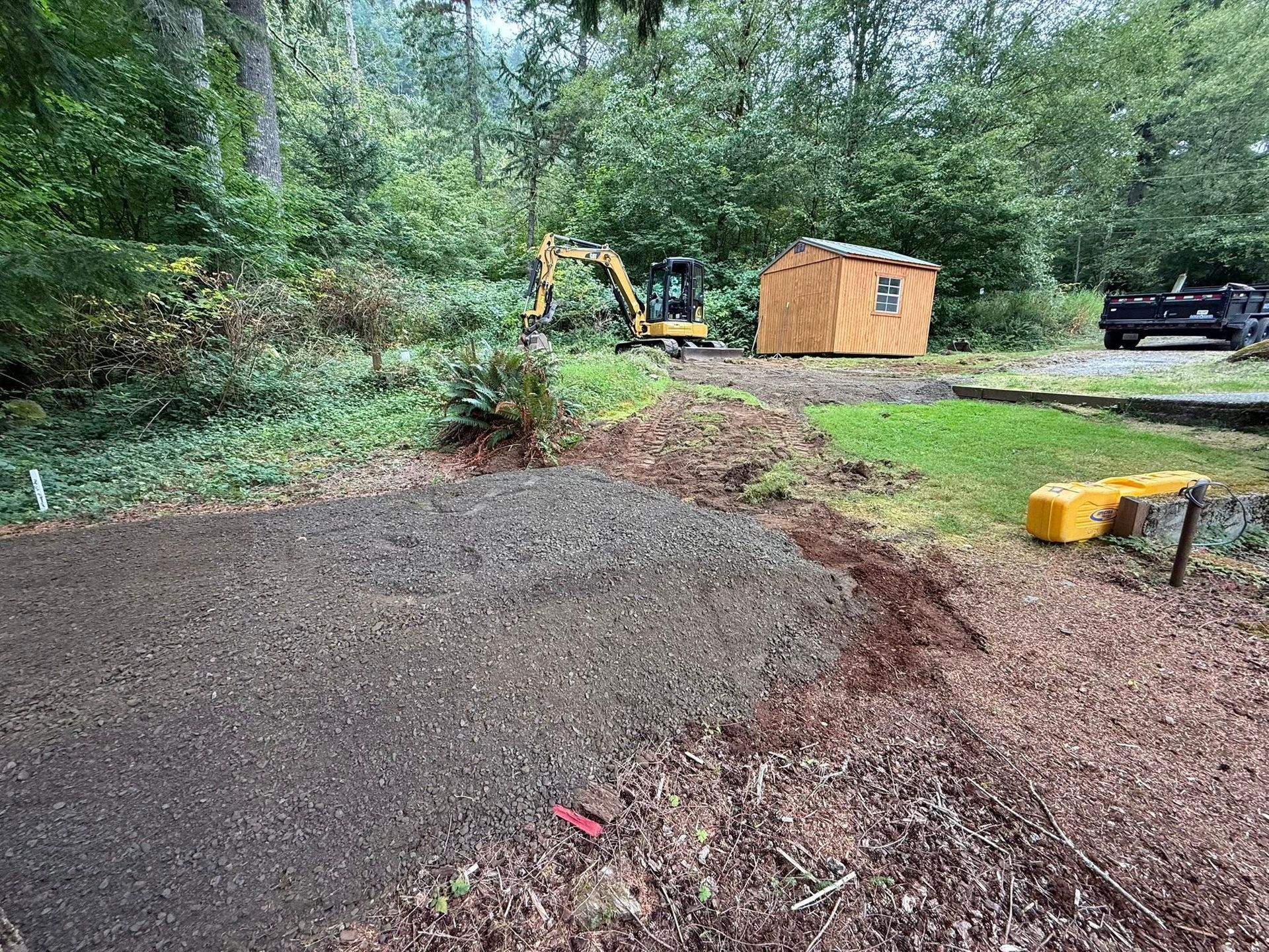 An excavator and a small shed sit in a wooded yard with an area of fresh gravel in the foreground.
