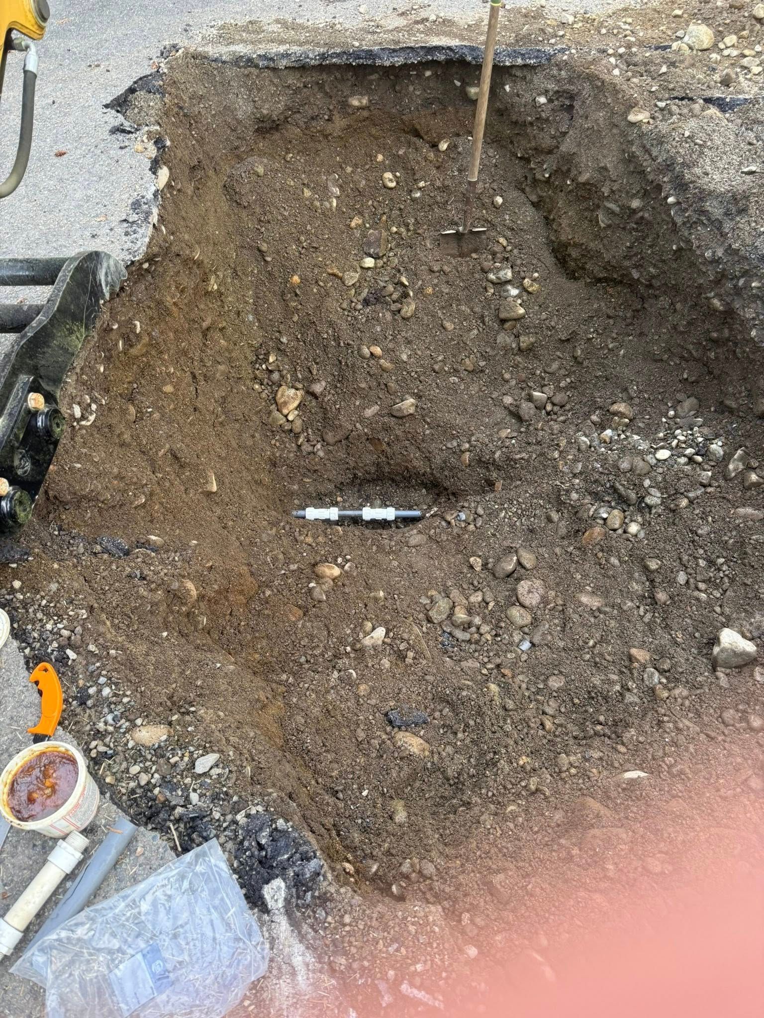 A rectangular trench dug into soil and asphalt, showing a horizontal black pipe and construction equipment nearby.