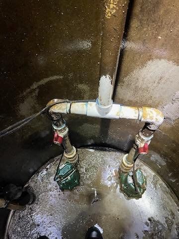 Two sump pumps connected by white PVC pipes to a central discharge pipe inside a concrete basin.