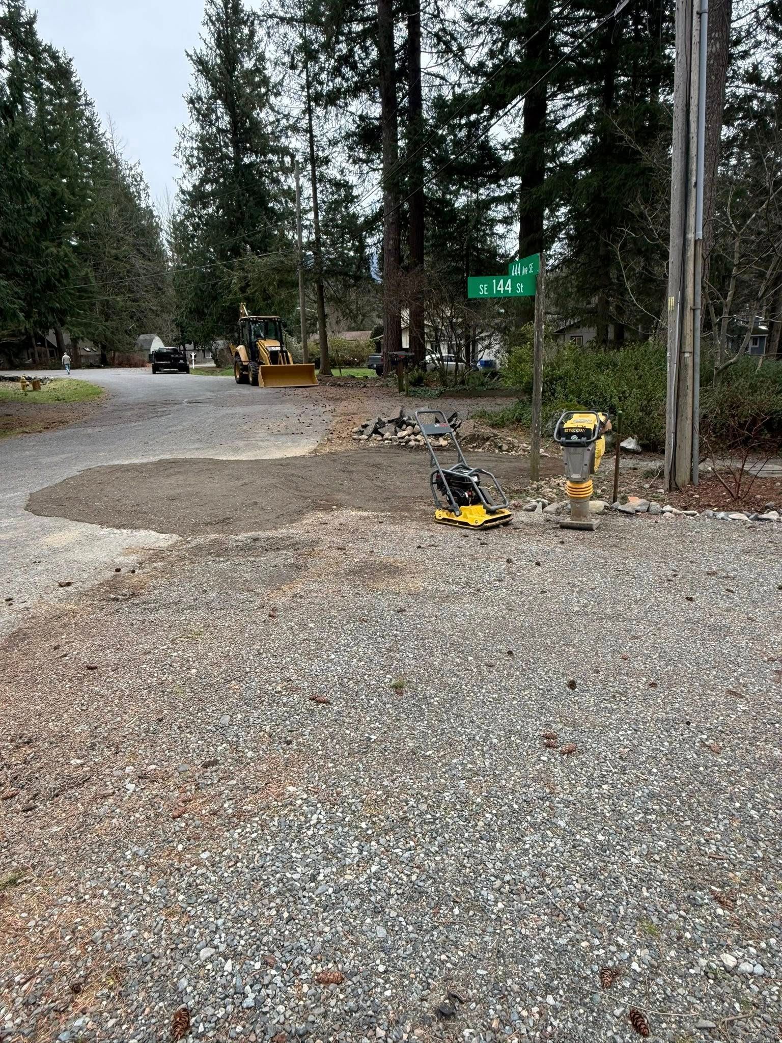 A construction worker with a yellow piece of equipment stands on a gravel road near a street sign and trees.