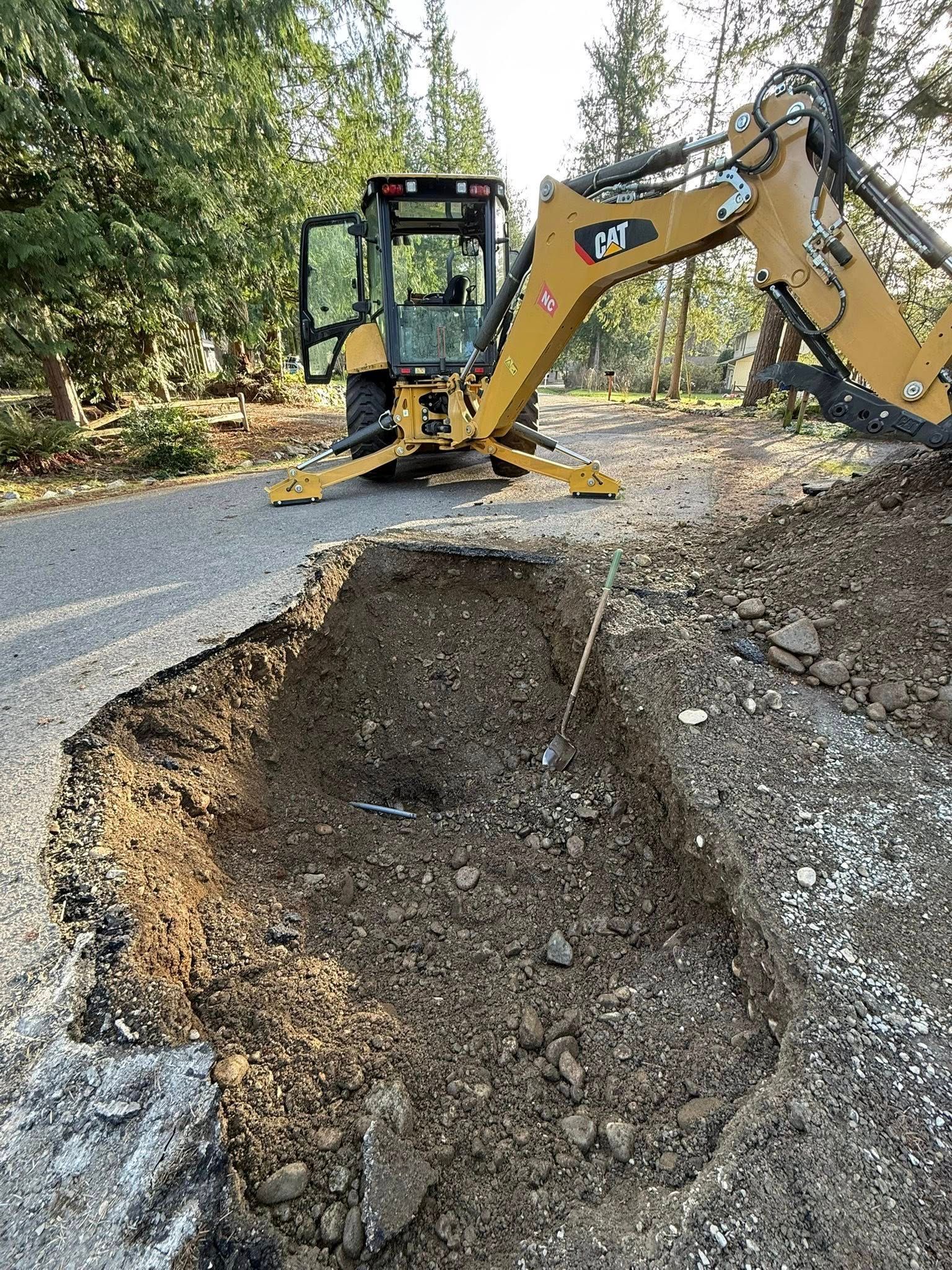 A yellow Caterpillar backhoe loader parked on a gravel road next to a large, freshly excavated trench.