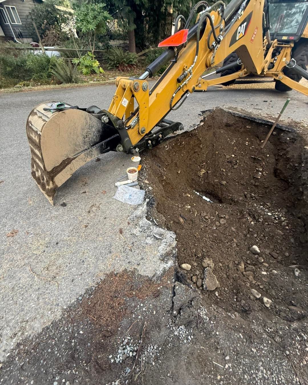 A yellow backhoe excavates a deep hole in an asphalt road, with a small plastic cup and debris sitting on the pavement.