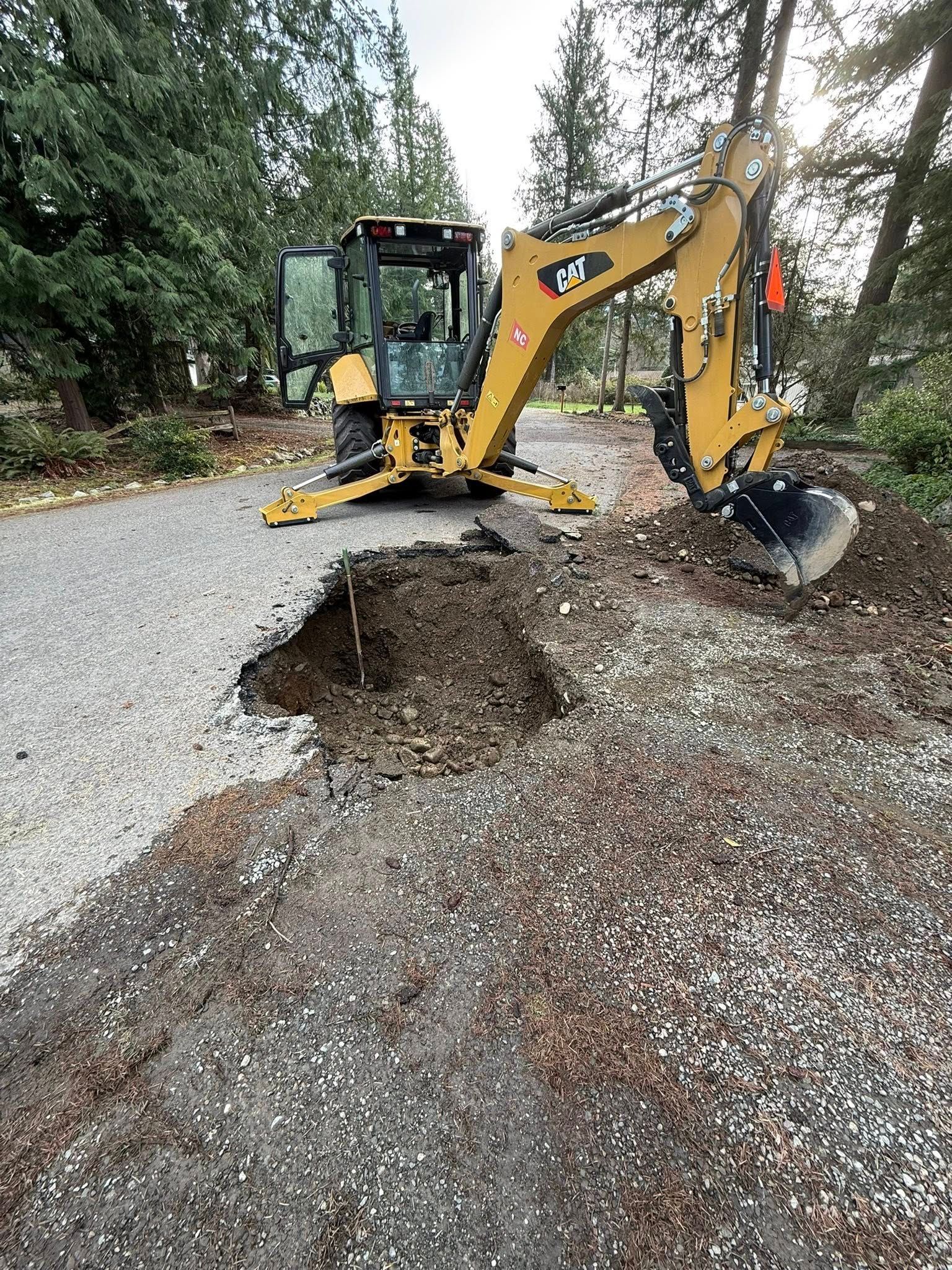 A yellow backhoe excavator is parked on a gravel road next to a freshly dug hole in the ground.