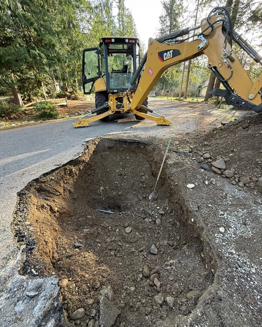 A yellow CAT backhoe parked on a paved road next to a freshly excavated trench in the dirt.