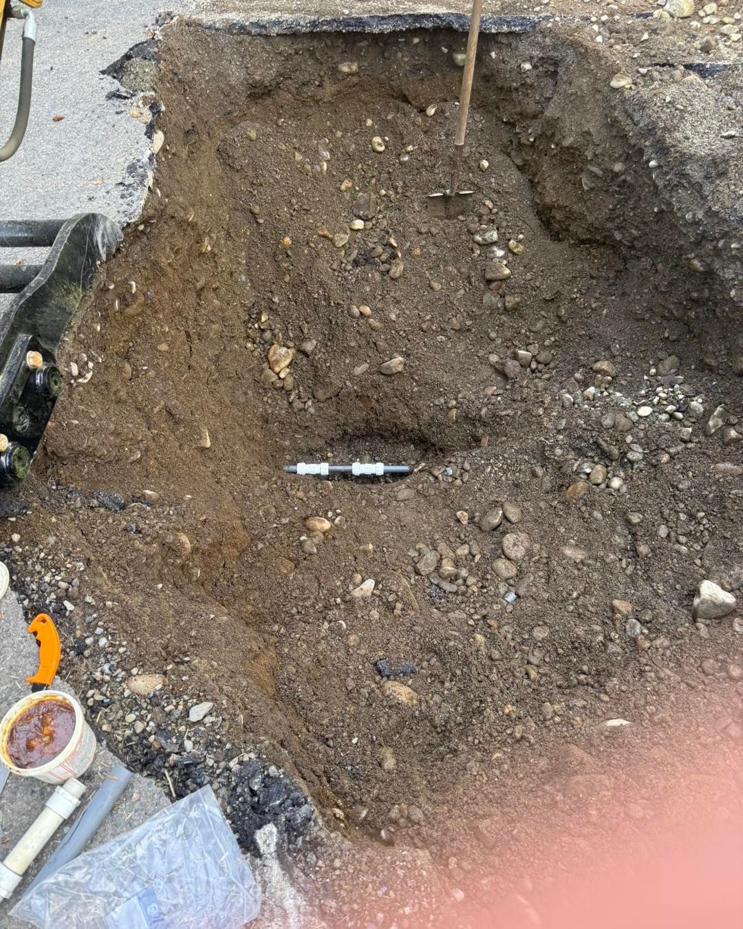 A small excavation in dirt reveals a dark pipe with a silver connector in the center, next to construction equipment.
