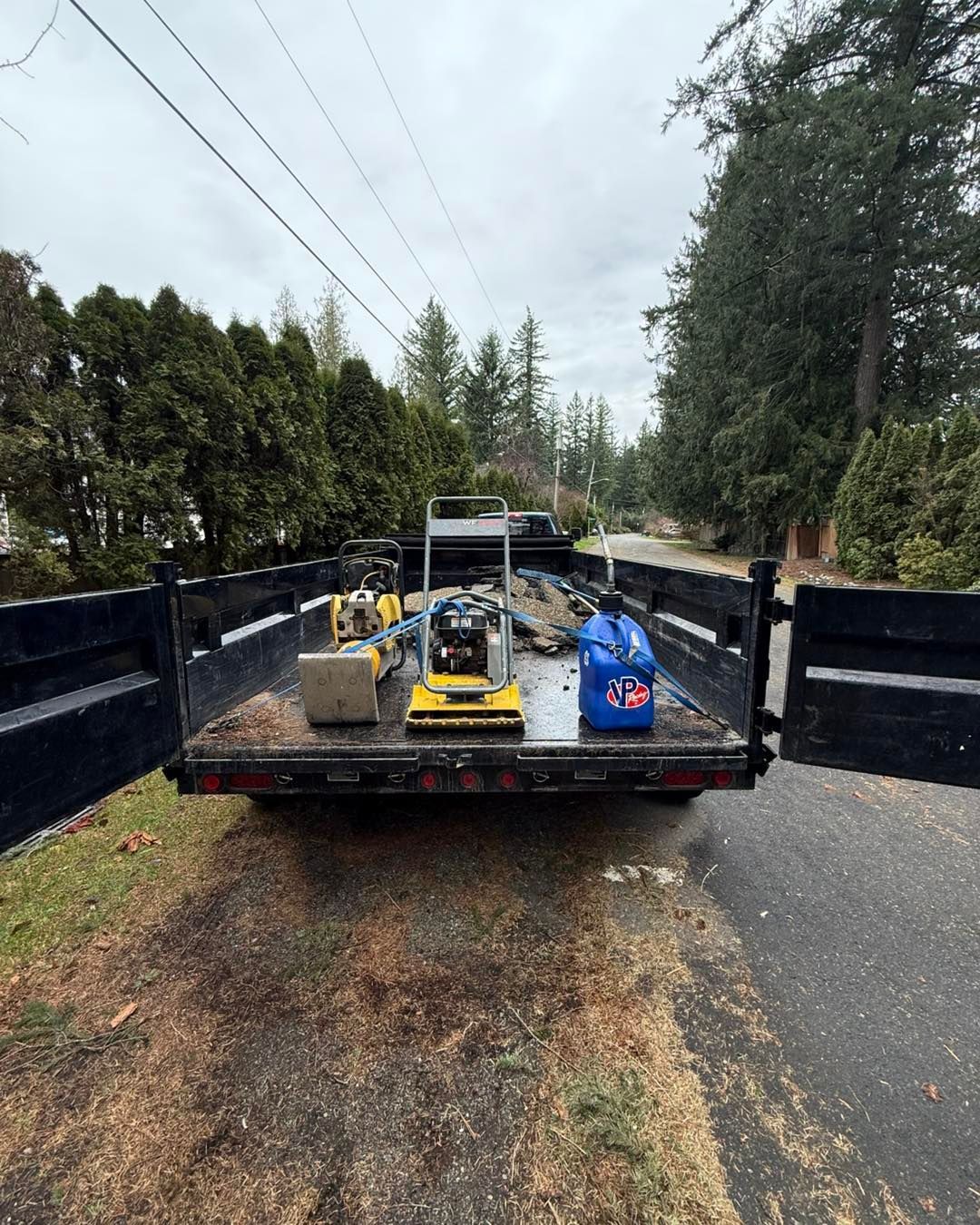 A flatbed trailer parked on a road holds two yellow plate compactors and a blue fuel container.