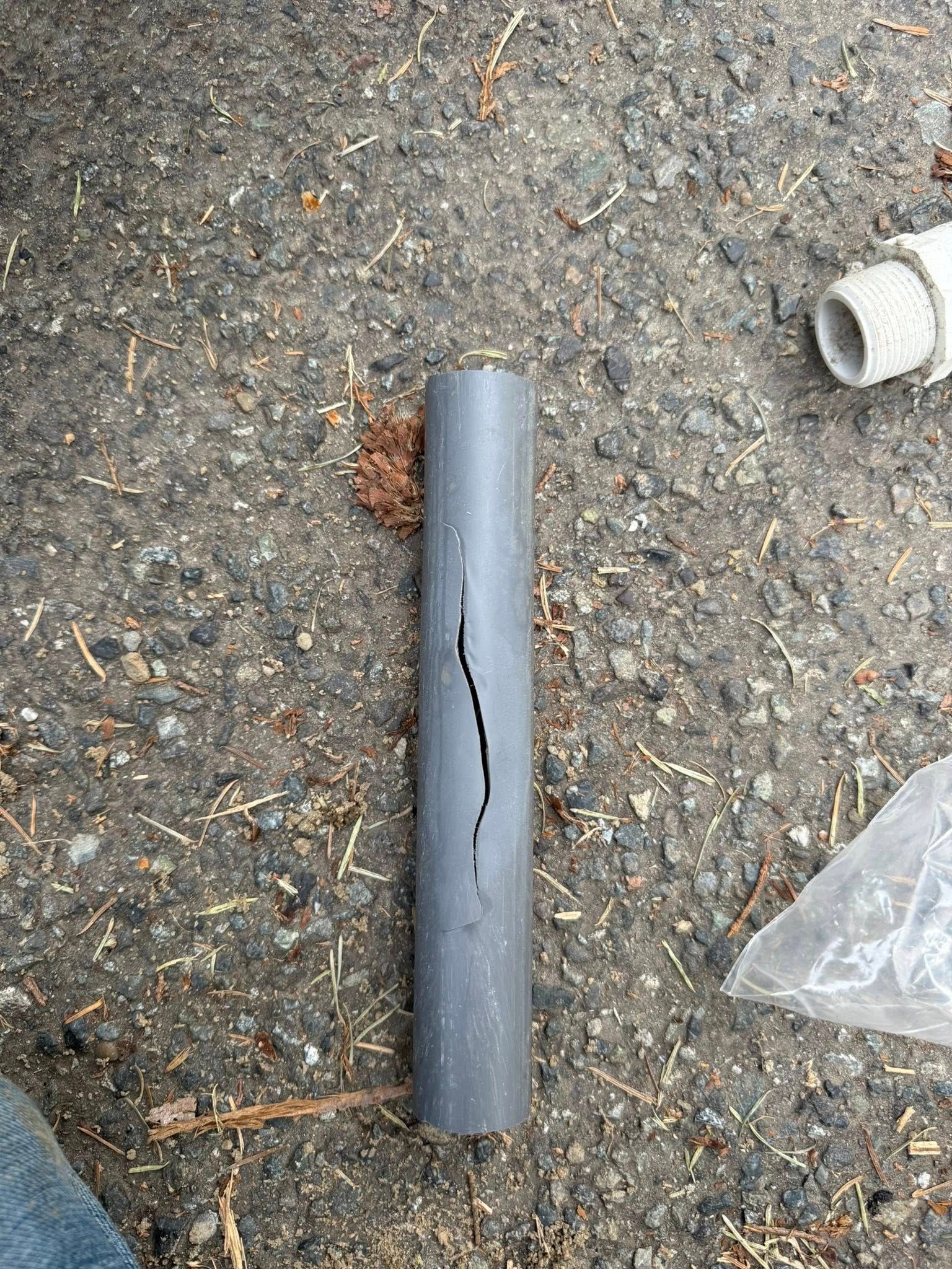 A gray plastic pipe lying on a rough, paved surface, featuring a long, vertical crack running down its center.