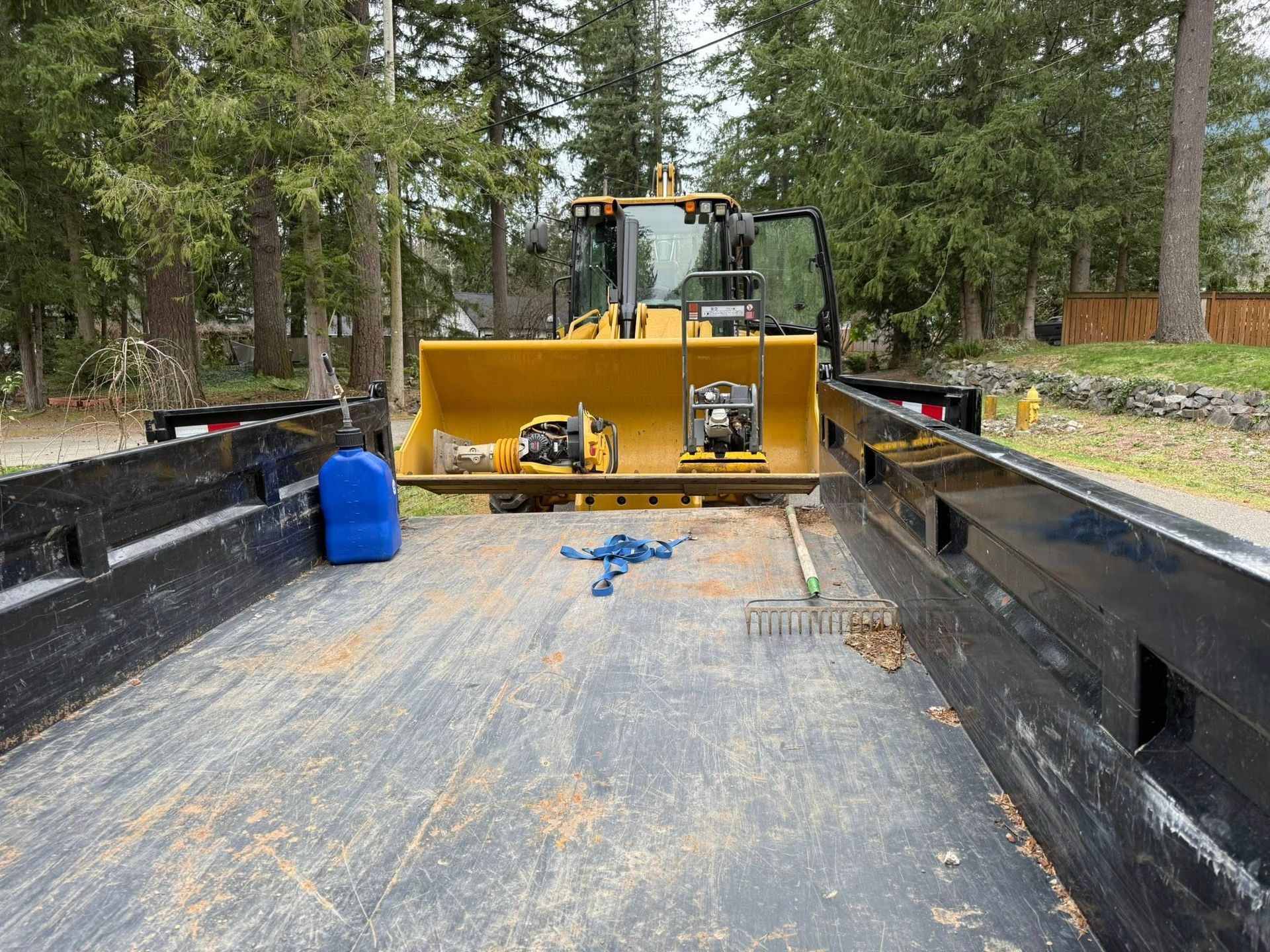 A yellow tractor bucket sits on a flatbed trailer containing a blue fuel jug, a rake, and tools in a wooded area.