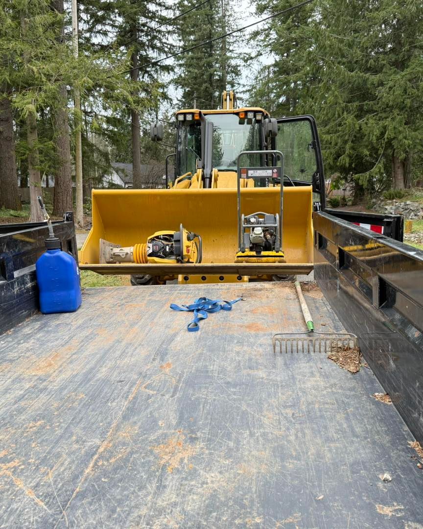 A yellow loader parked in the back of a truck, with a tamper and a compactor in its bucket, plus a rake on the floor.